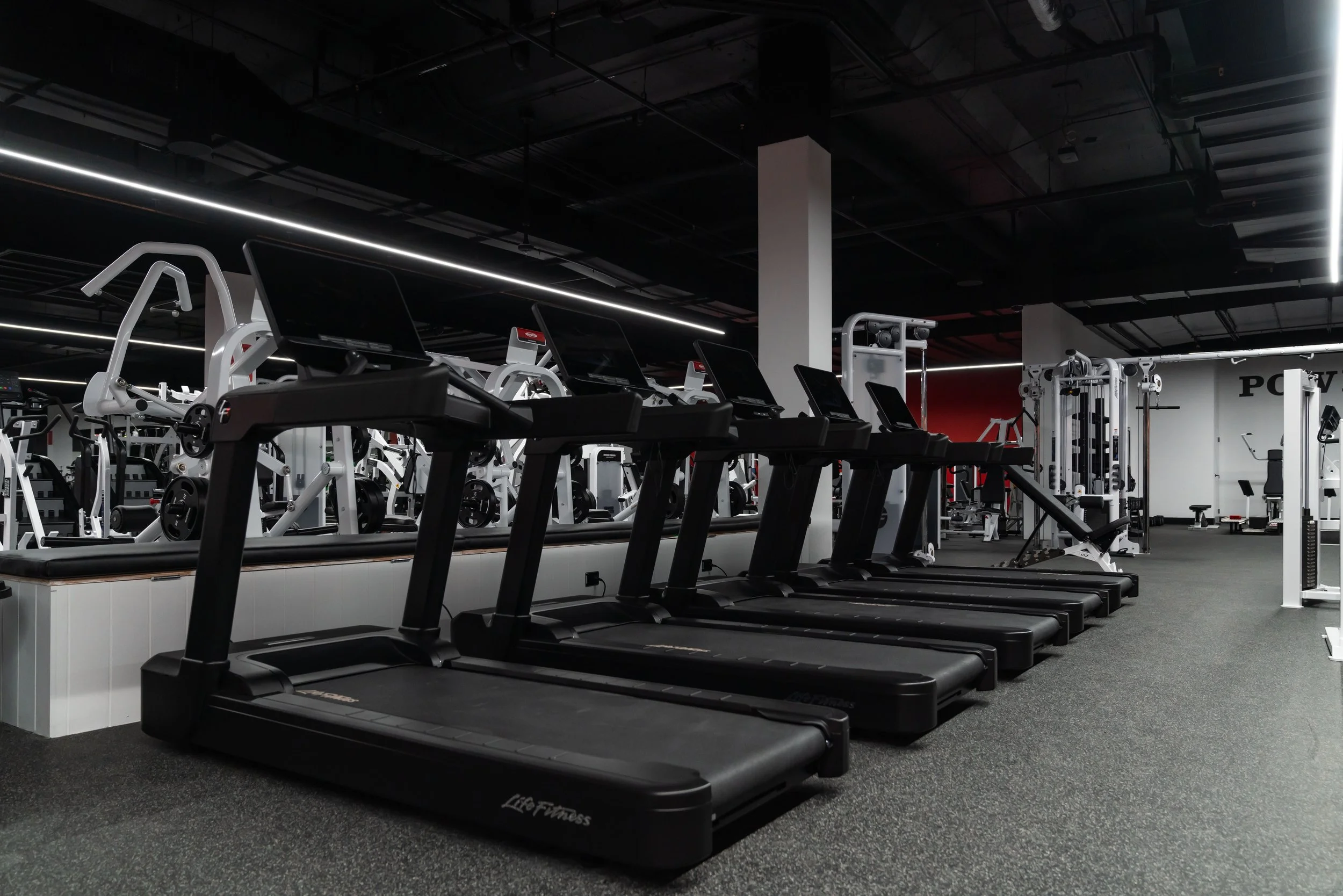 Row of black treadmills in a modern gym with various exercise equipment in the background.
