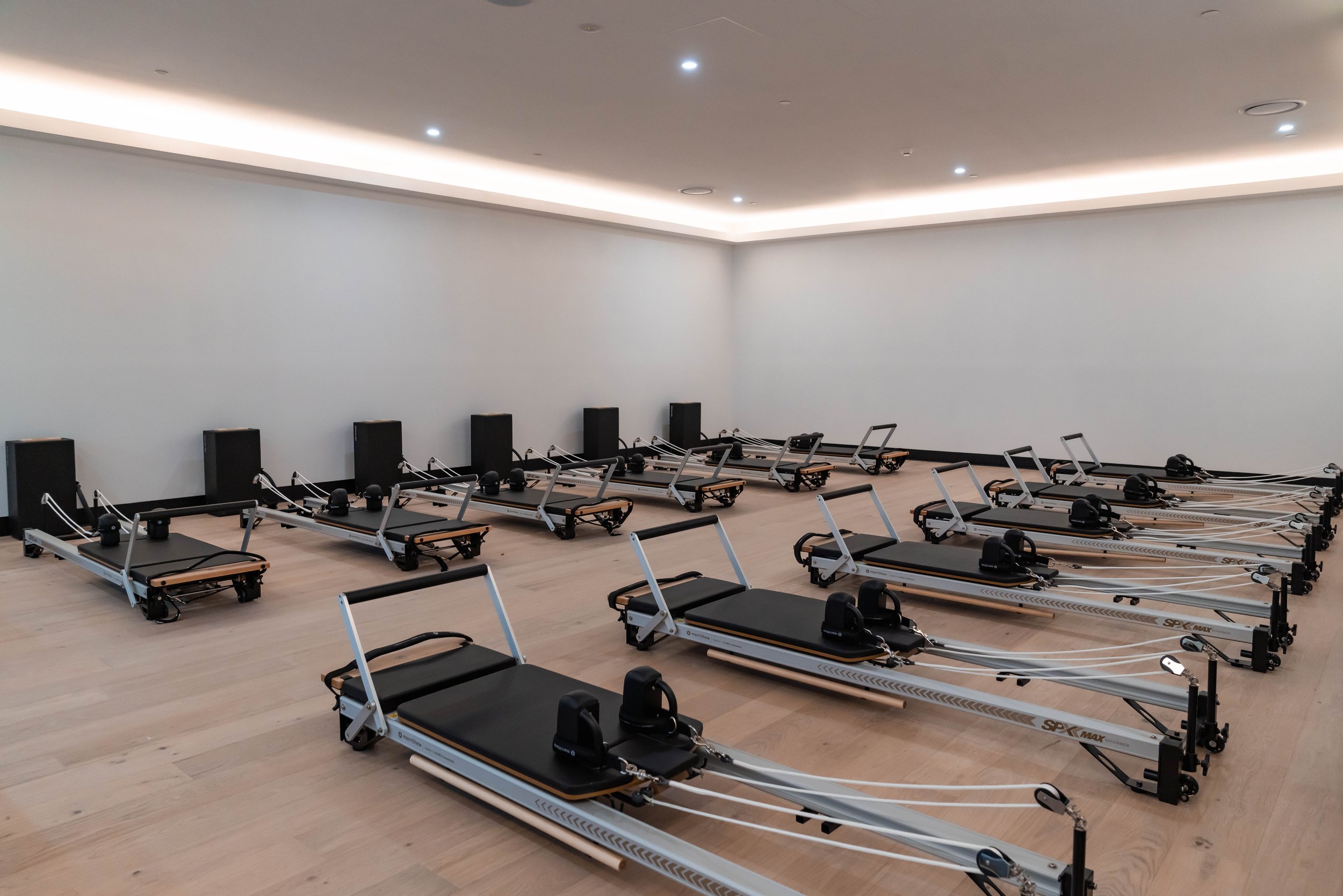 A spacious Pilates studio with multiple reformer machines arranged in rows on a light wood floor, white walls, and recessed ceiling lights.