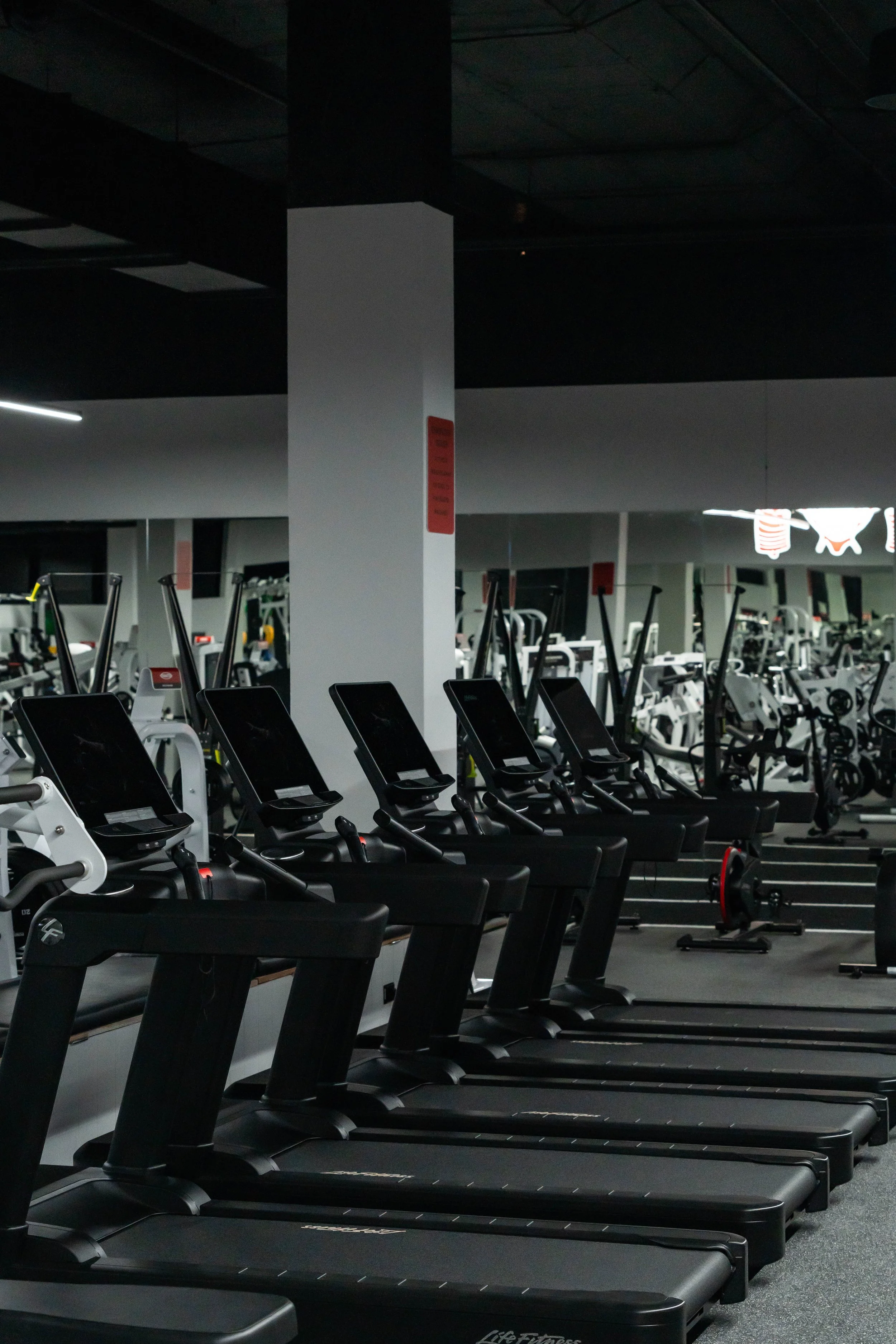 Row of black treadmills in a dimly lit gym with weight machines and mirrors in the background.