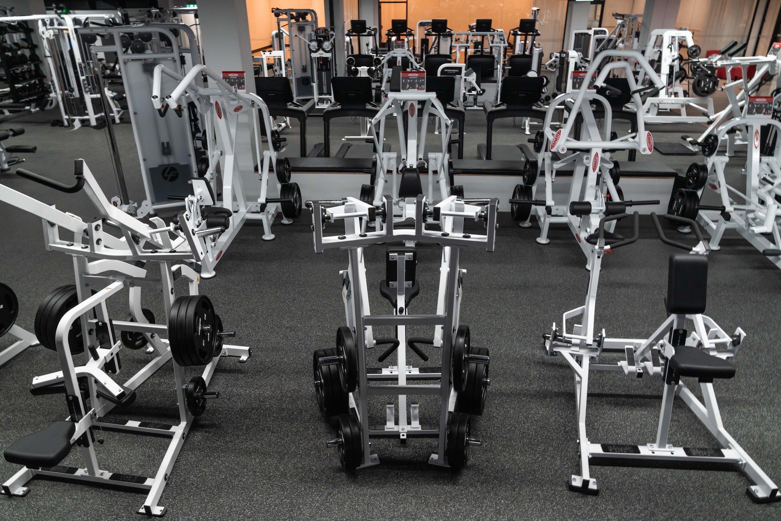 gym with white weightlifting machines and black chairs, arranged in rows on dark rubber flooring.