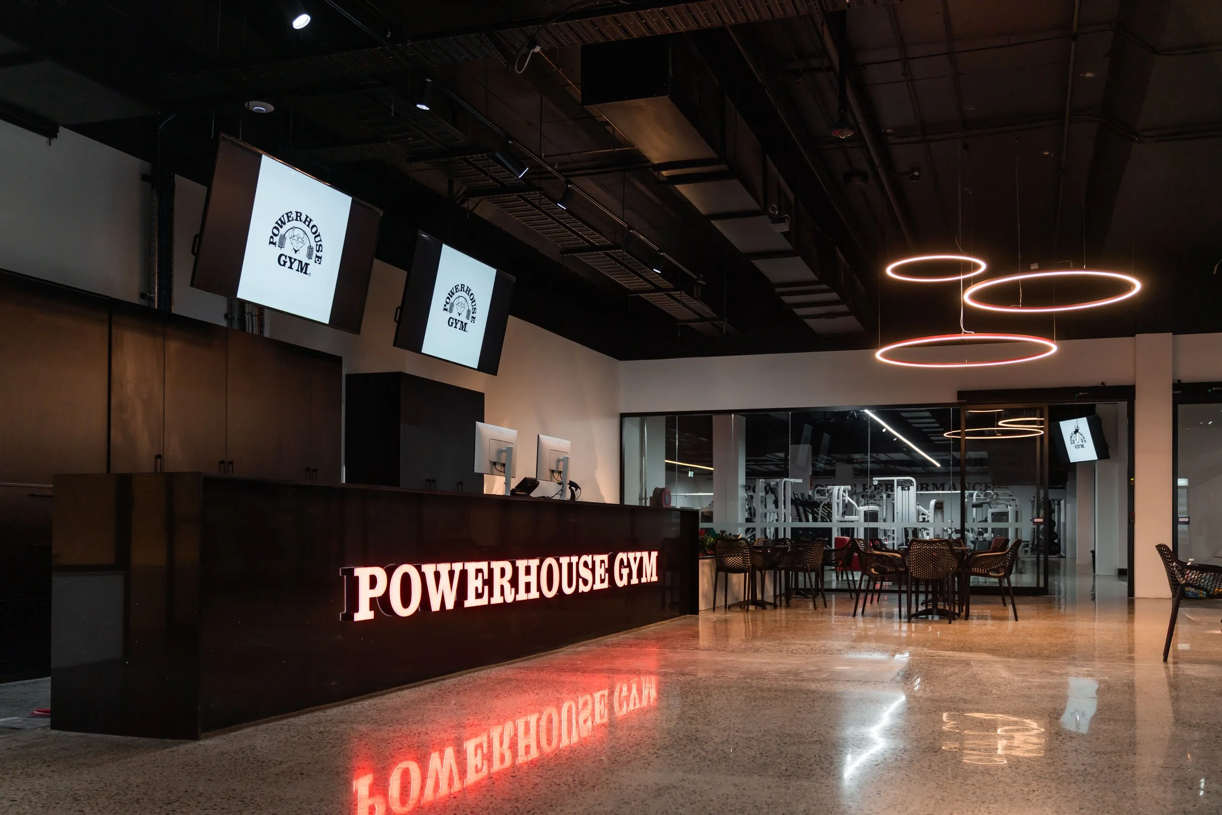 Reception area of Powerhouse Gym with a reception desk, illuminated sign, multiple monitors, seating area, and gym equipment visible in the background.
