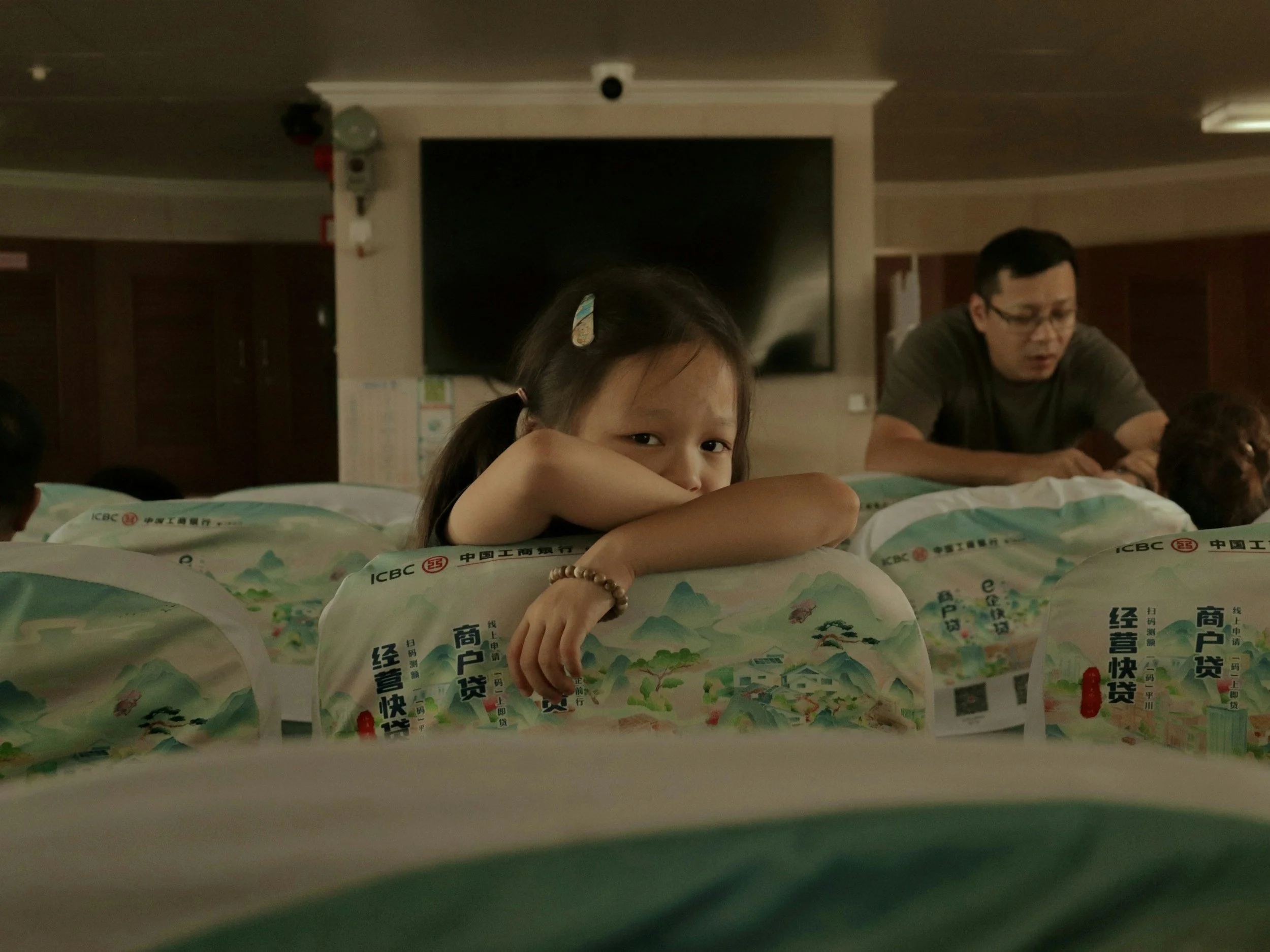 A girl looks sad and pensive while a parent is in the background.