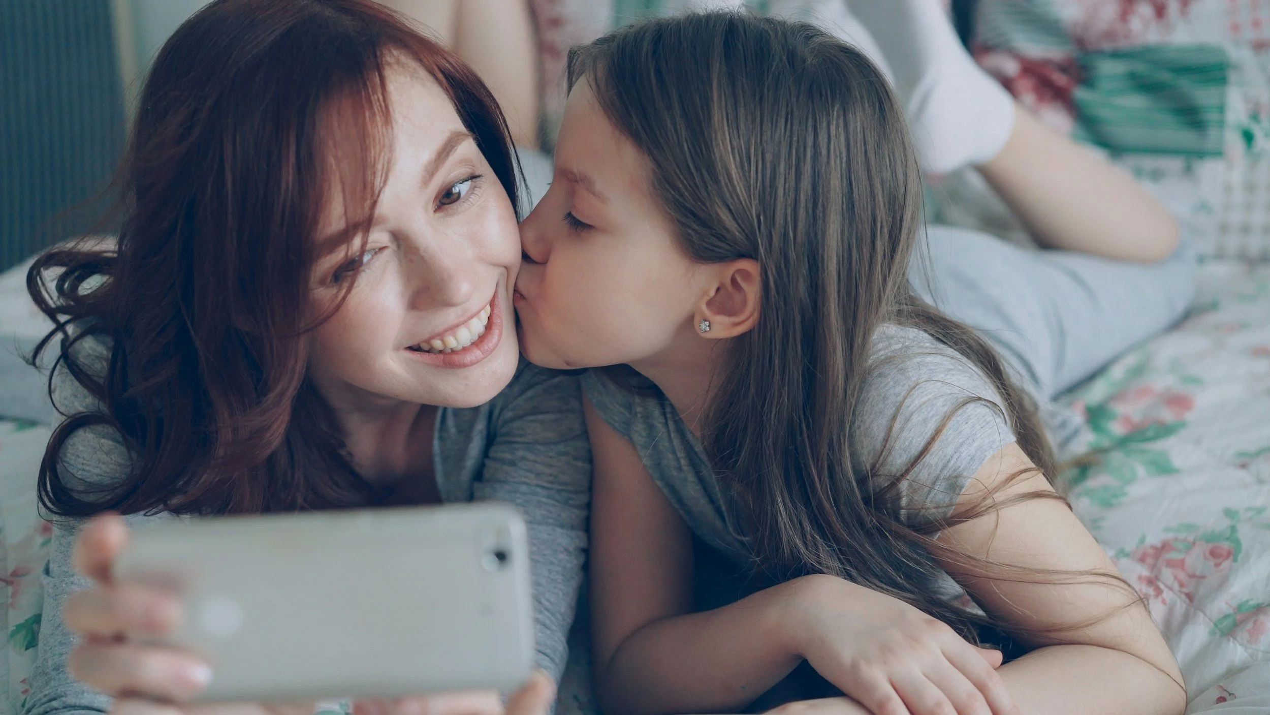 A mother holds her phone while the daughter kisses her on the cheek.