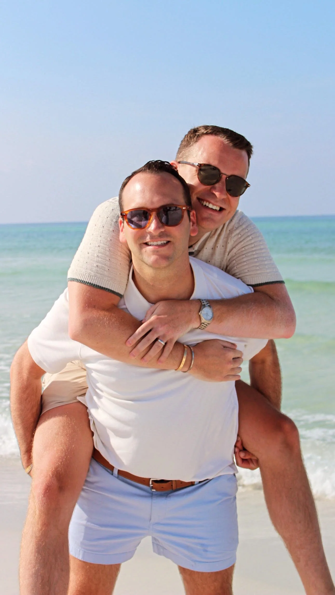 Two smiling men wearing sunglasses on a beach, one sitting on the other's back, with ocean and blue sky in the background.