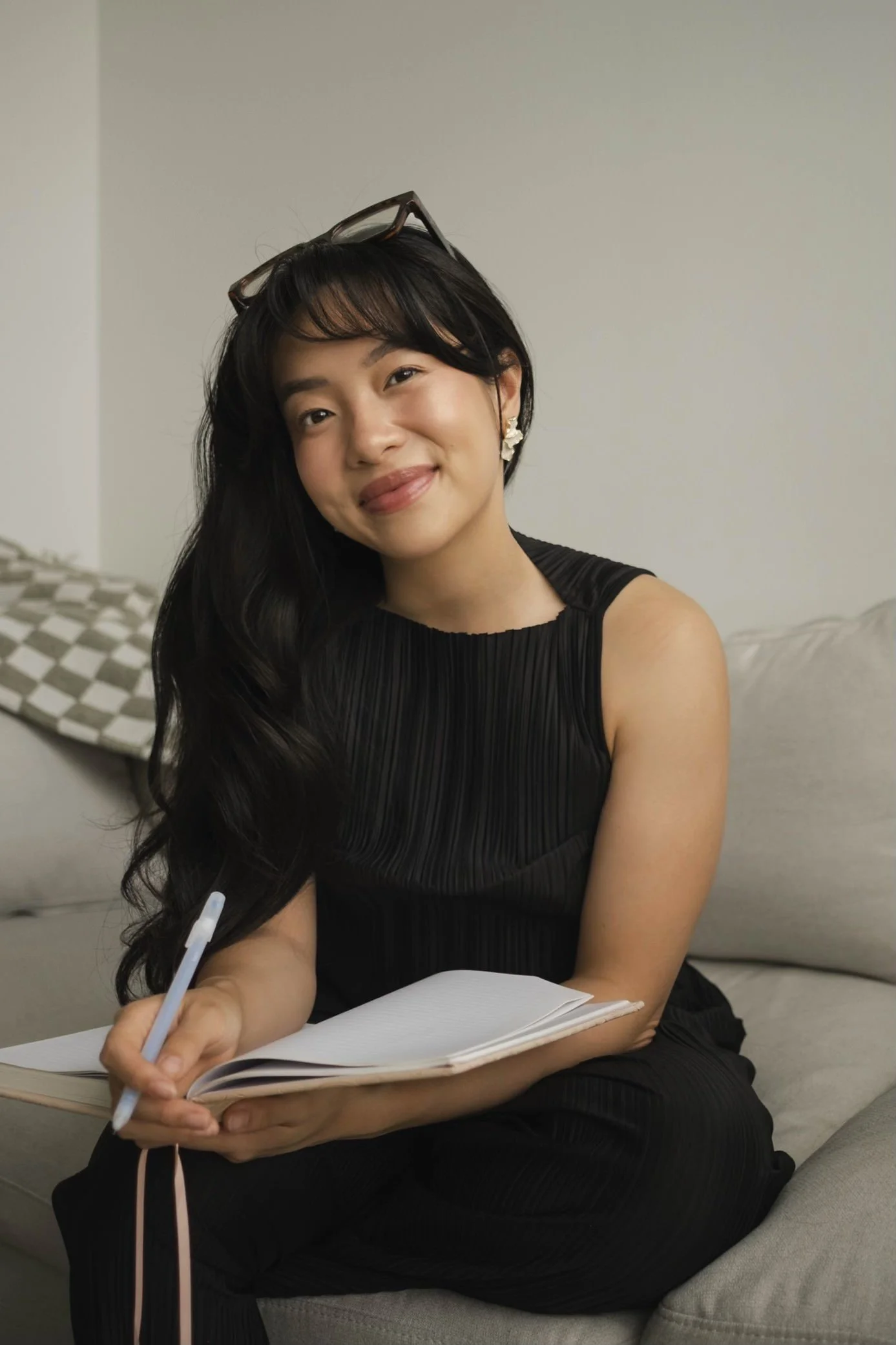A therapist woman sitting on a beige couch, holding an open notebook and pen, smiling at the camera, with glasses resting on her head and earrings.