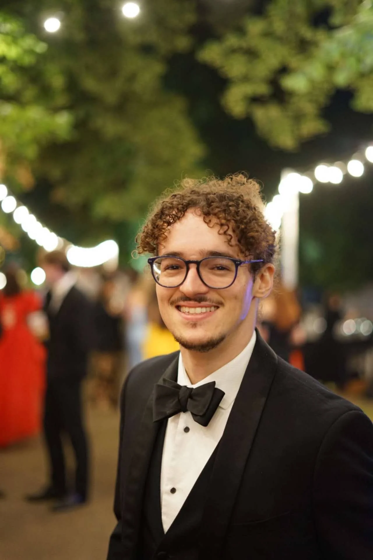 A smiling young man with curly hair and glasses wearing a tuxedo with a bow tie at an outdoor evening event, with blurred lights and people in the background.