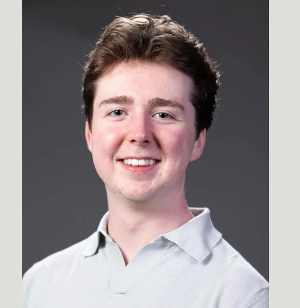 A young man with brown hair, blue eyes, and fair skin, smiling, wearing a light-colored collared shirt, against a dark gray background.