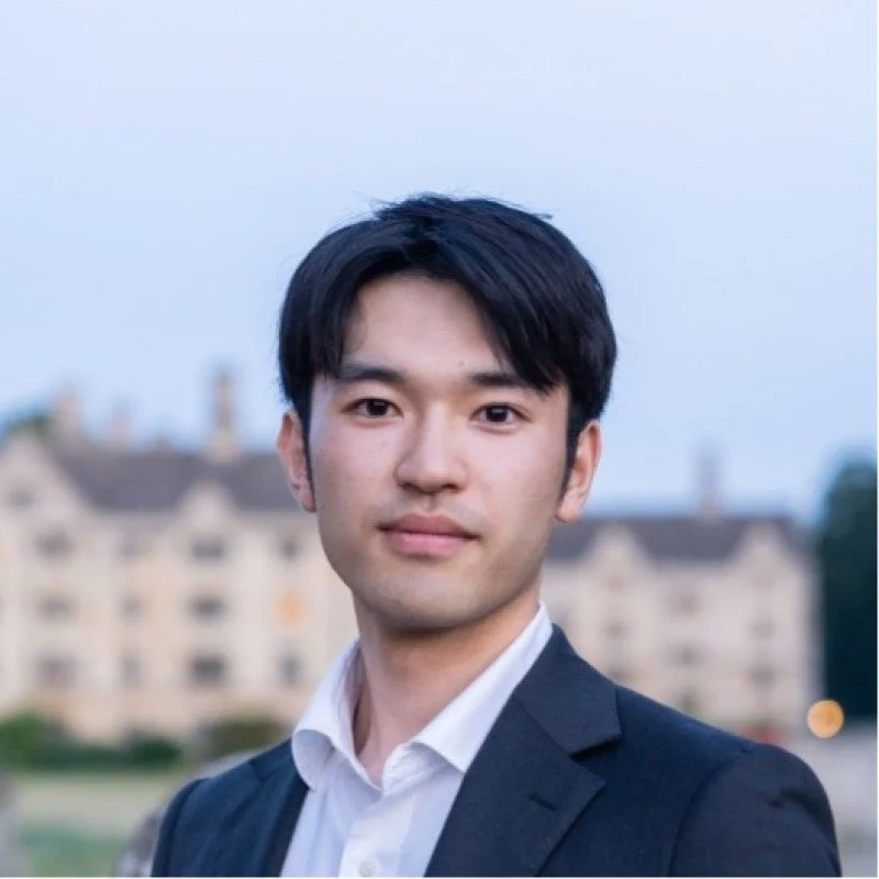 A young man wearing a dark suit jacket and white dress shirt outdoors, with blurred buildings and sky in the background.