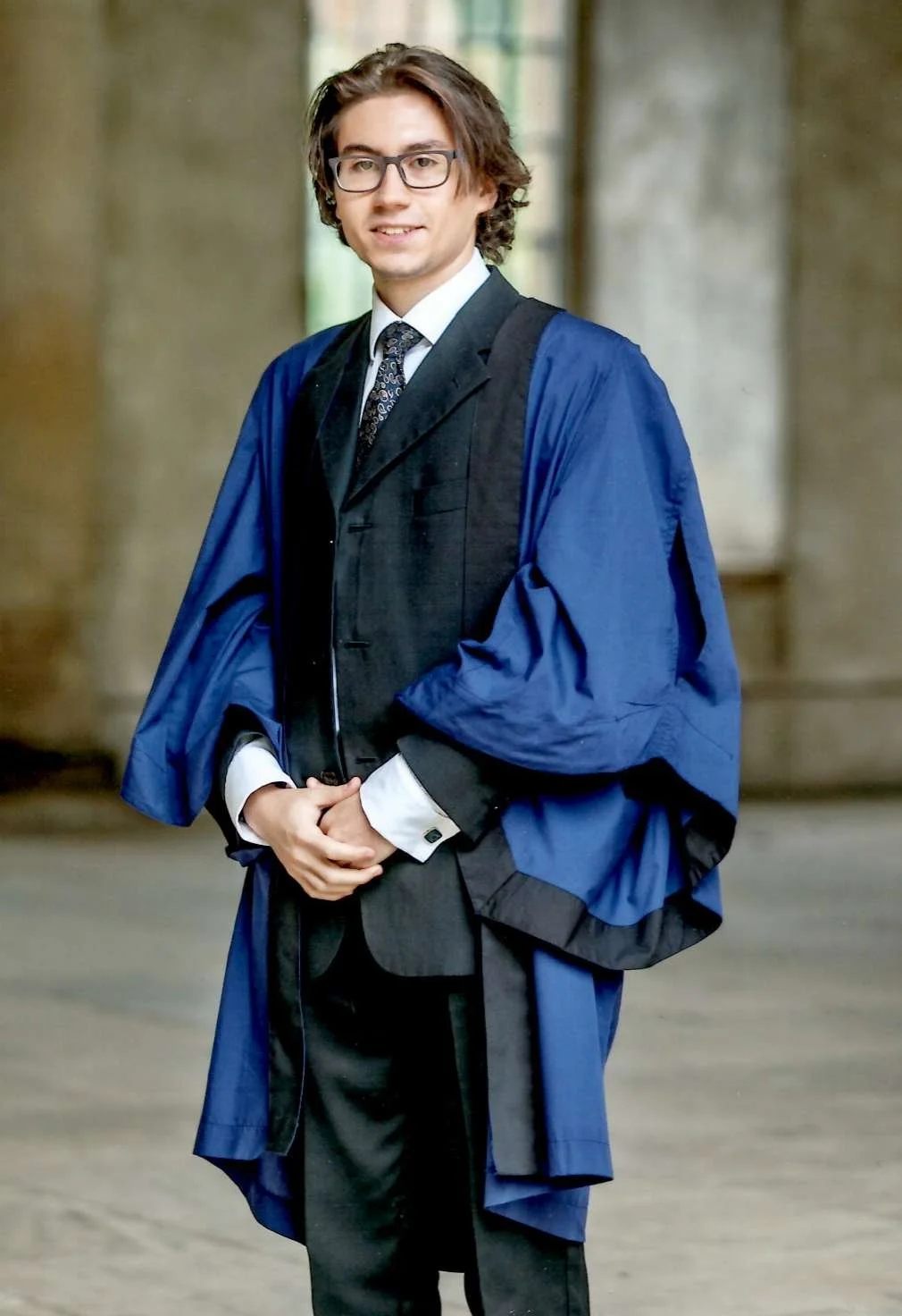 A young man wearing glasses in a graduation gown and cap, standing indoors with a blurred background.
