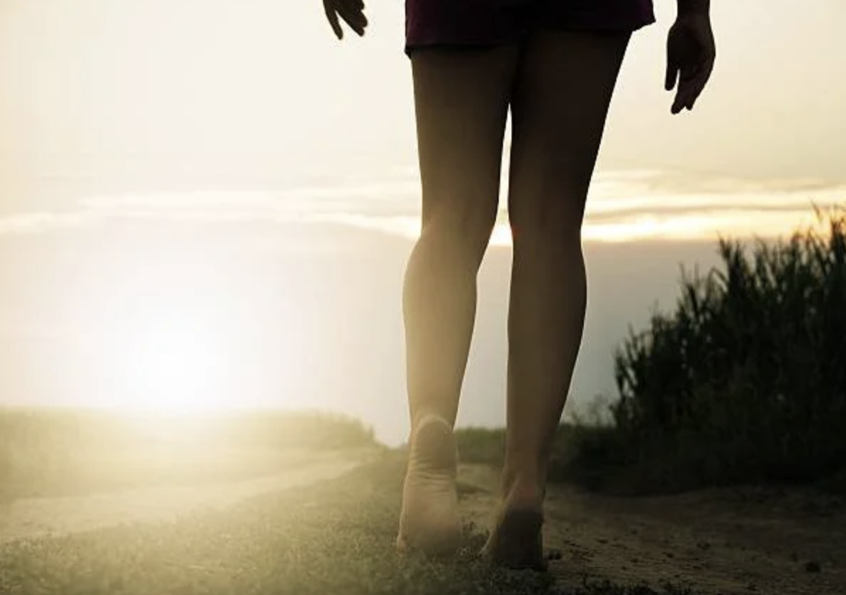Person walking barefoot on a sandy path at sunset, wearing dark clothing and gloves.