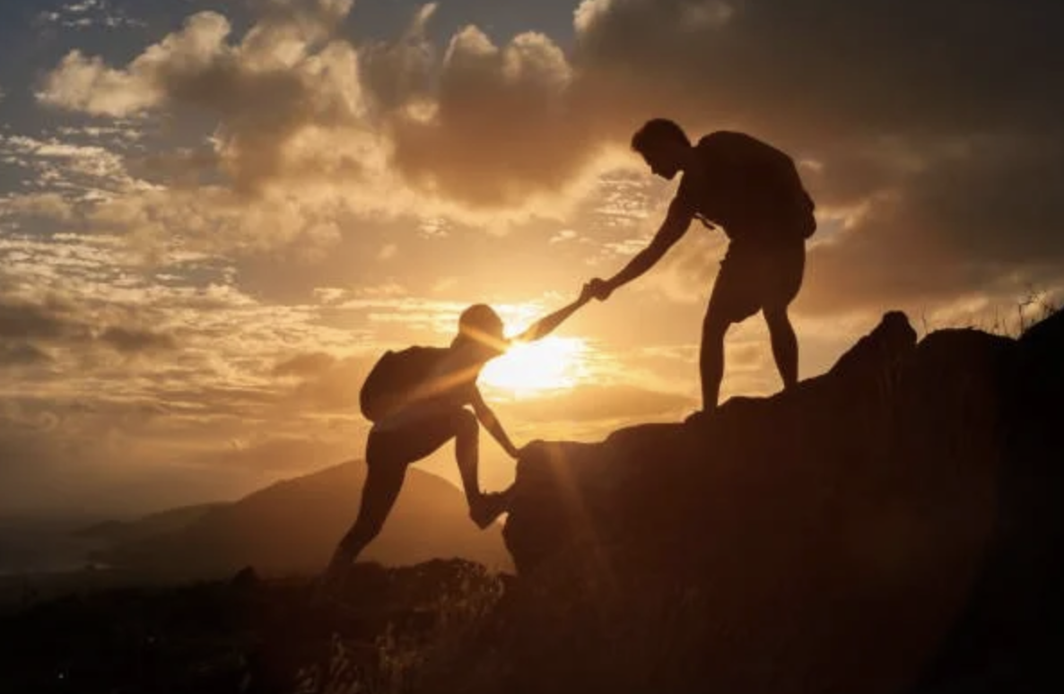 Silhouettes of two people on a mountain during sunset, with one helping the other to climb up.
