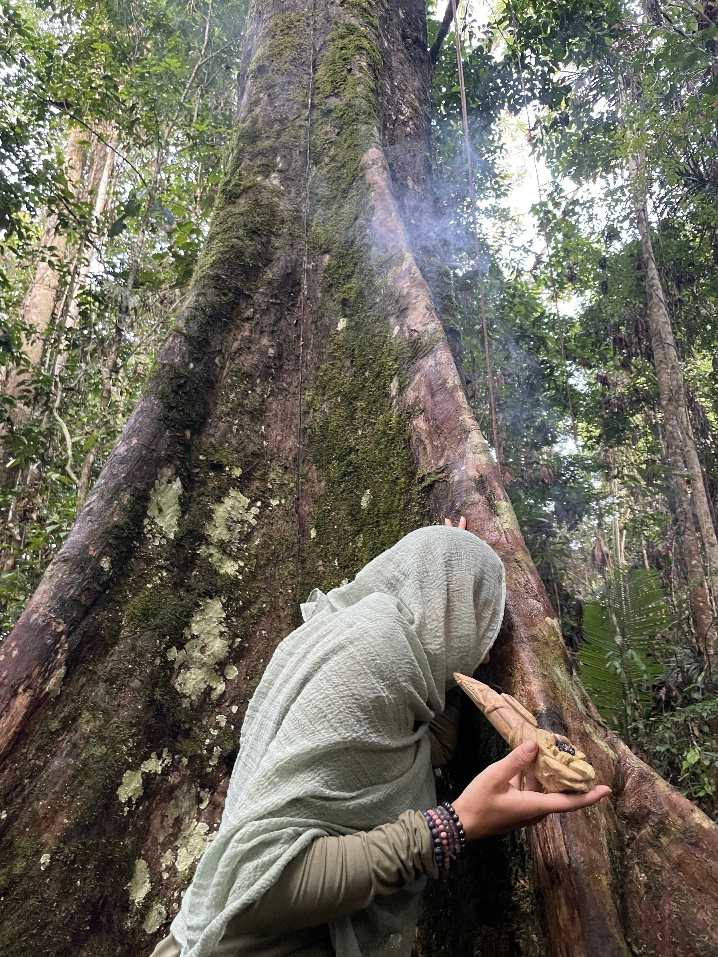 Person in a headscarf hugging a large tree in a dense forest, holding a carved wooden object.