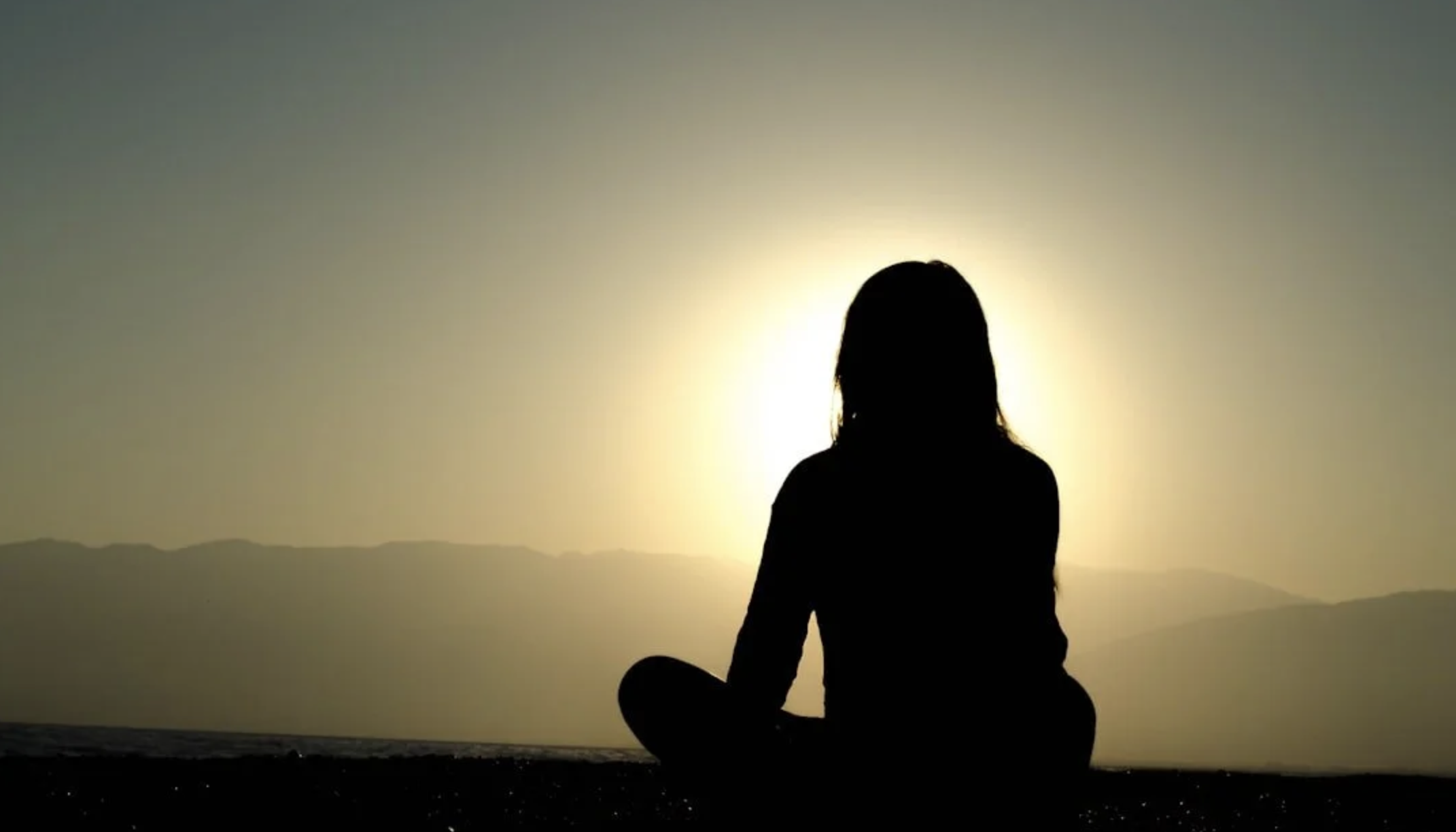Silhouette of a person sitting in meditation on the ground at sunset or sunrise, with mountains in the background.