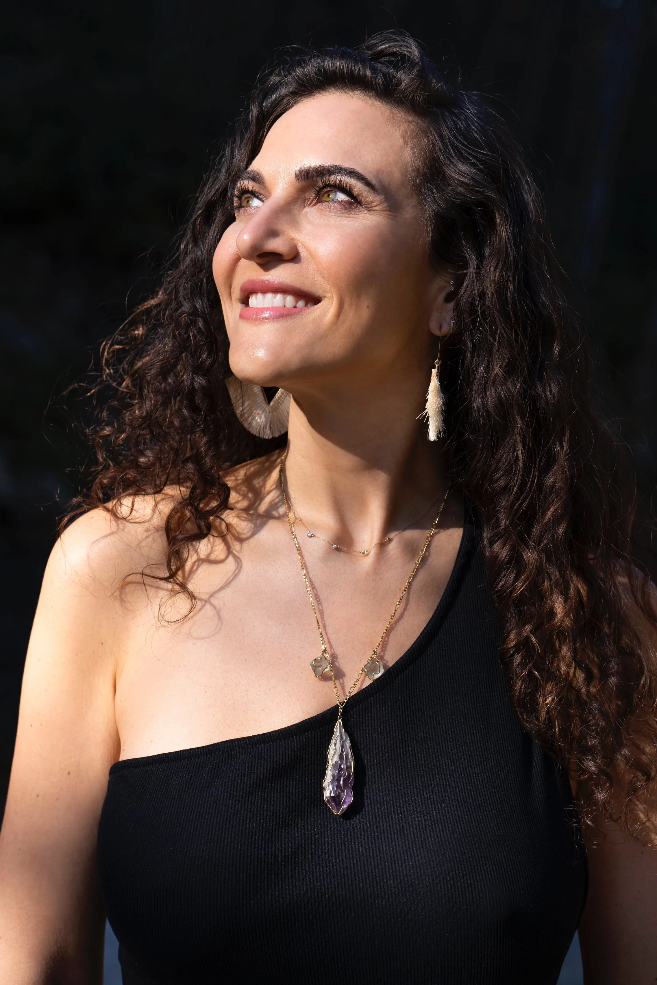 A woman with long curly dark hair smiling, wearing a black sleeveless top and decorative jewelry, looking slightly upward.