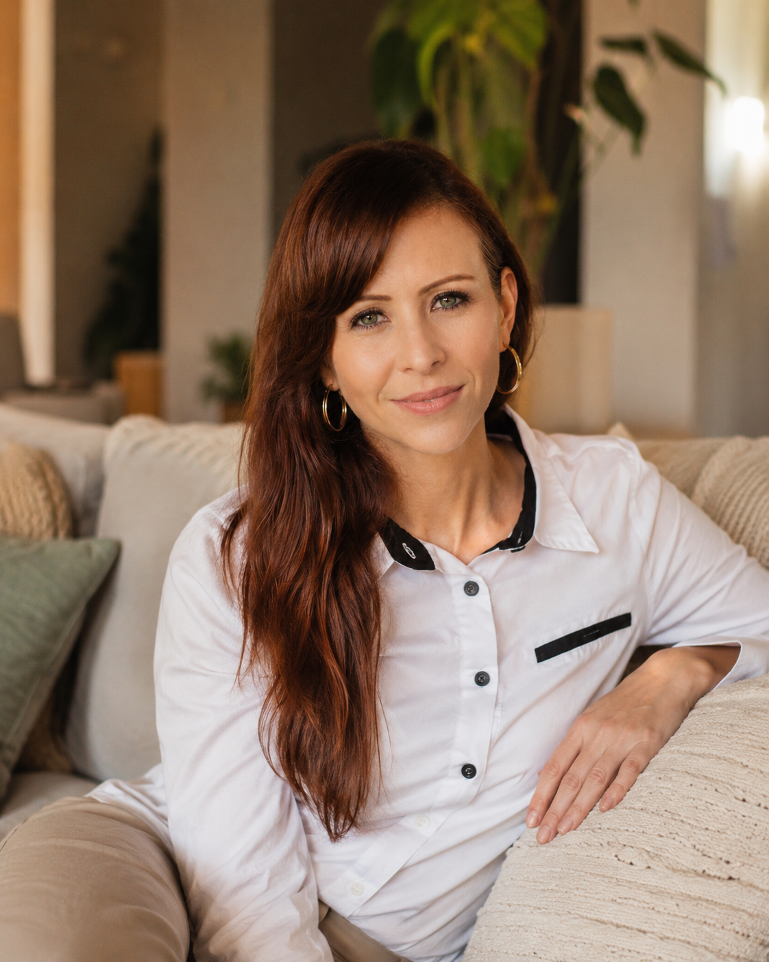 A woman with long, reddish-brown hair and hoop earrings sitting on a cream-colored couch in a living room.