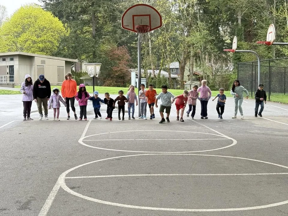 Young children and adults standing in a line on a basketball court, holding hands, preparing to jump or hop together outdoors with trees and basketball hoops in the background.