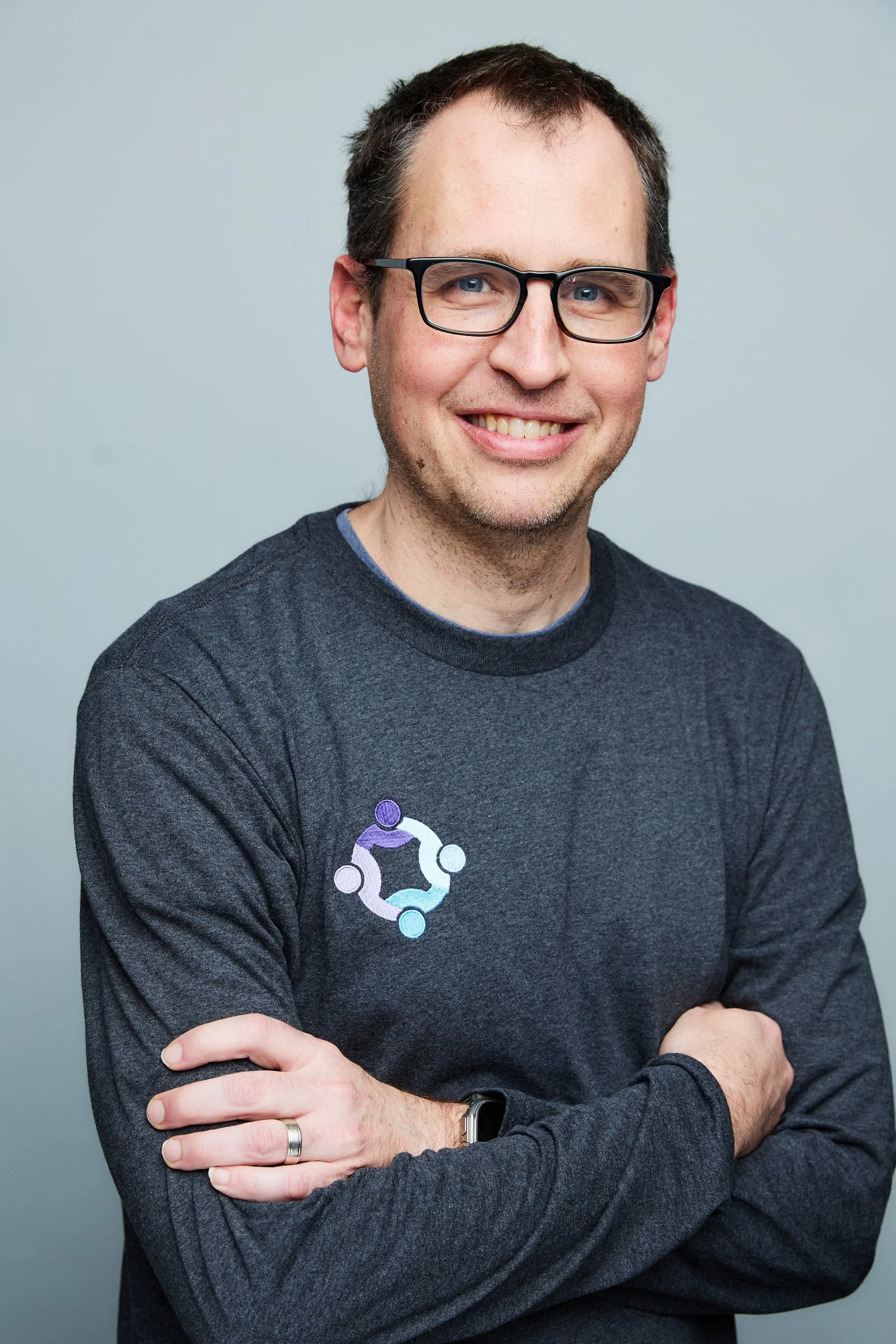 A smiling man with glasses and short dark hair, wearing a dark gray long-sleeve shirt with a logo on the chest, standing with arms crossed against a plain light gray background.