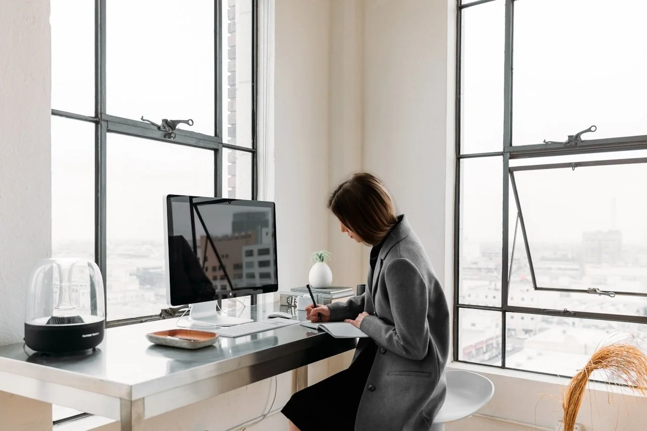 Woman in gray blazer sitting at a desk near large windows, writing in a notebook, with a computer monitor, potted plant, and small appliance on the desk.