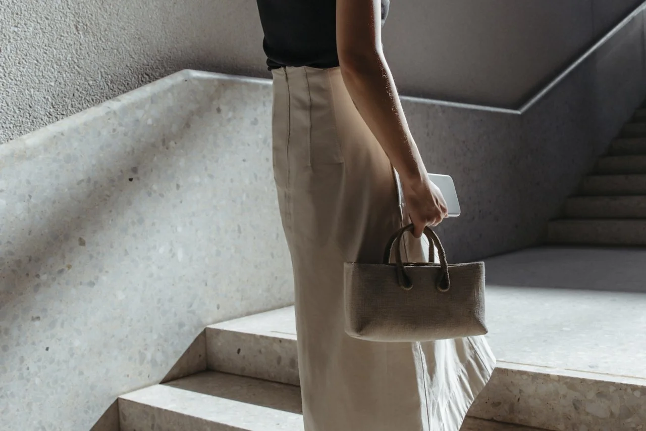 A person wearing a black top and beige skirt holding a beige handbag and a phone, standing on a concrete staircase indoors.