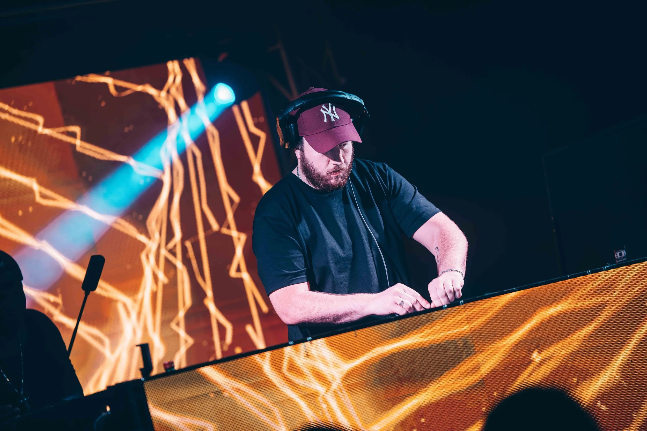 A DJ wearing a maroon baseball cap and headphones performs behind a digital DJ booth with a bright, lightning-like digital display in the background.