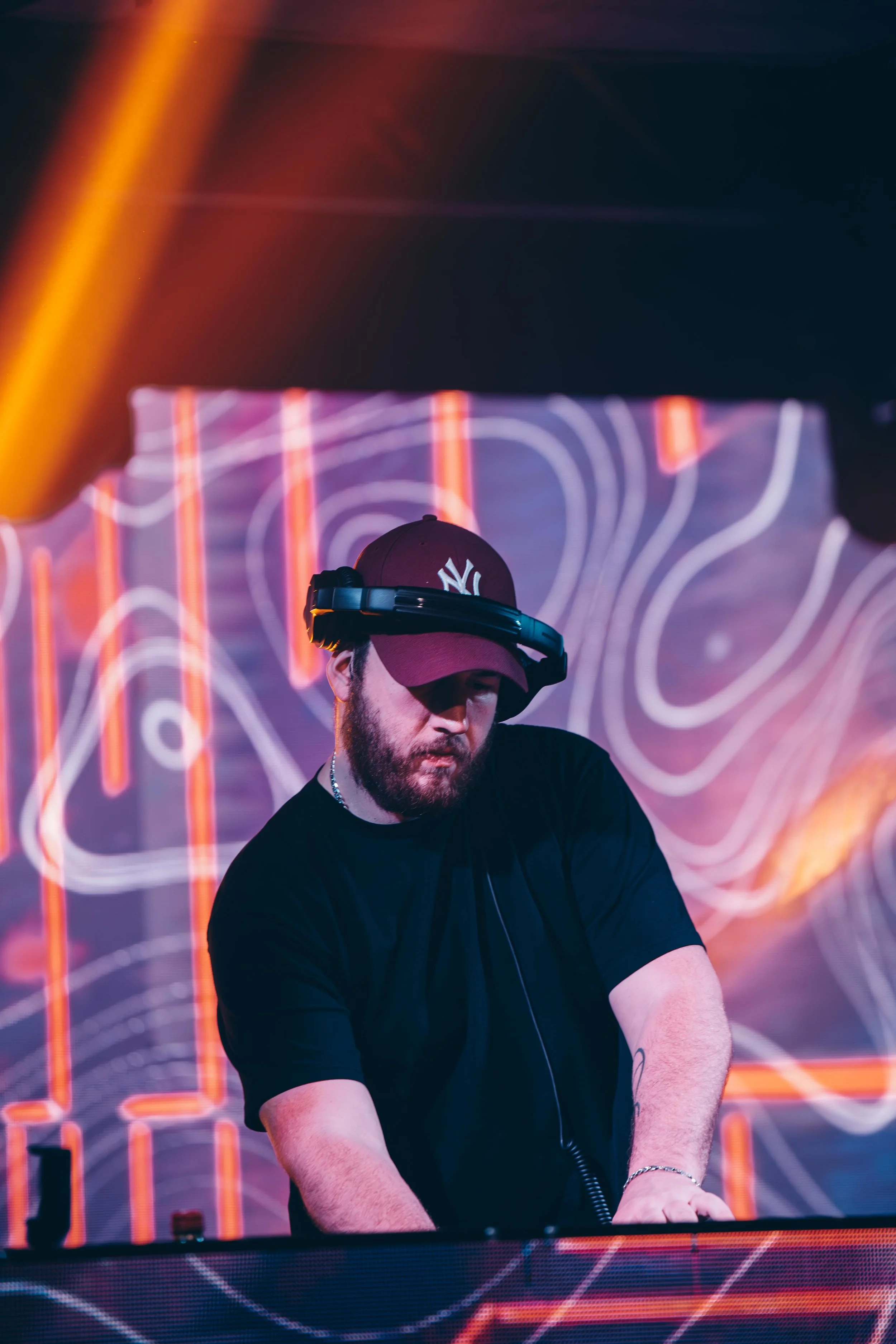A DJ wearing a black t-shirt, a New York Yankees cap, and headphones stands behind a DJ booth with neon lights in the background.