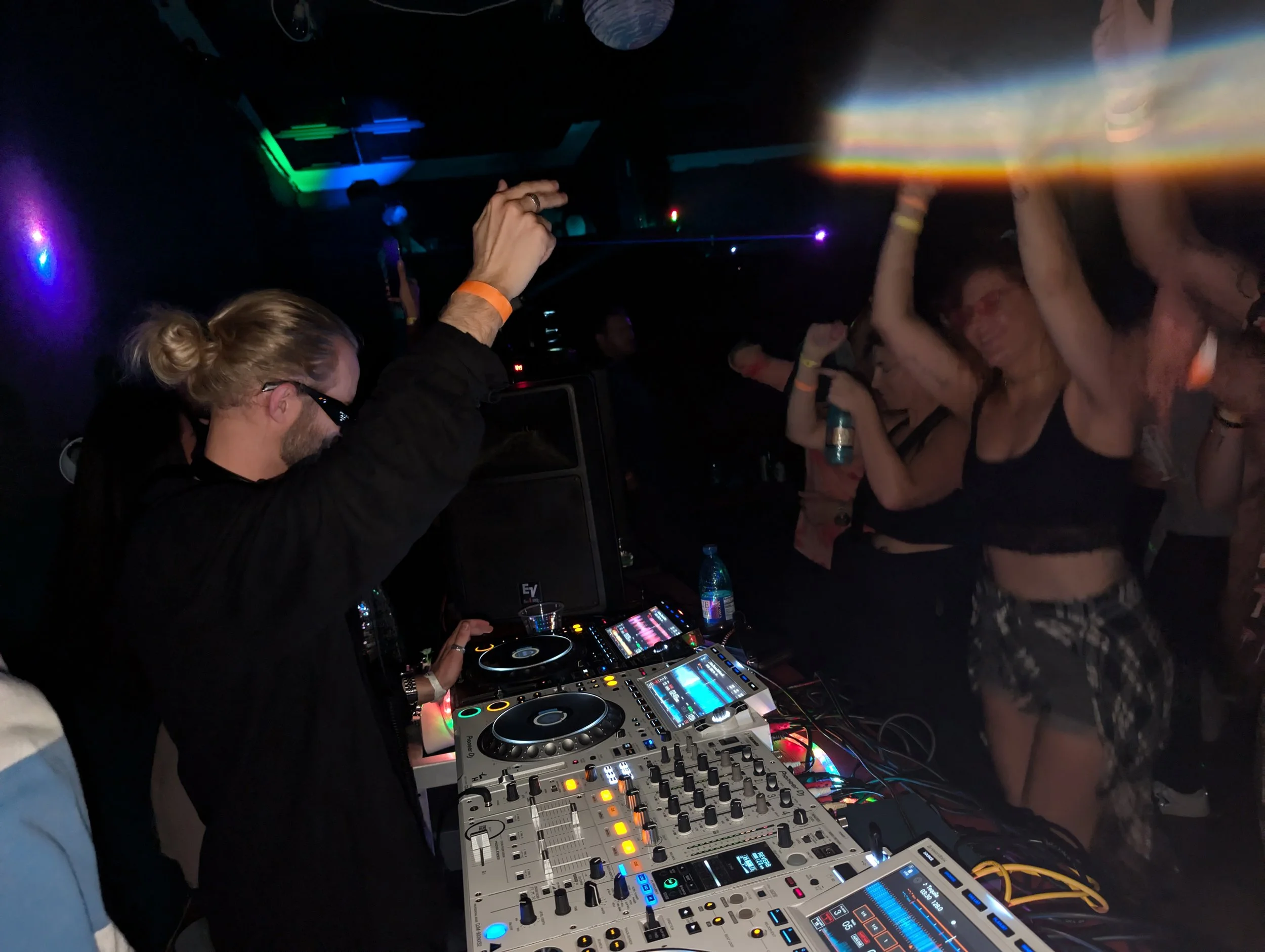 People dancing at a nightclub with a DJ playing electronic music, with colorful lights and a dark atmosphere.
