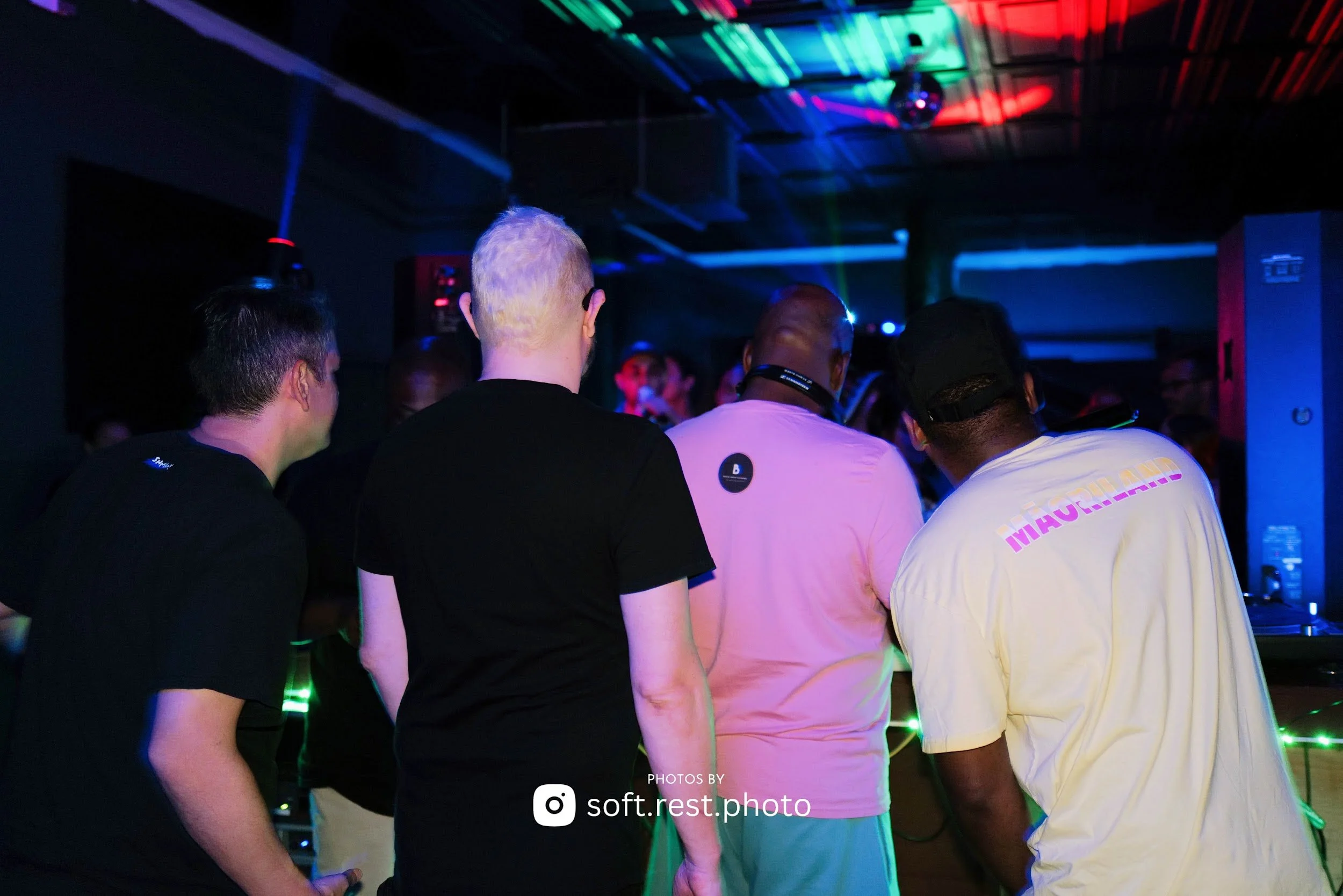 Group of five people standing closely together in a dimly lit room with colorful lights, possibly at a party or nightclub.