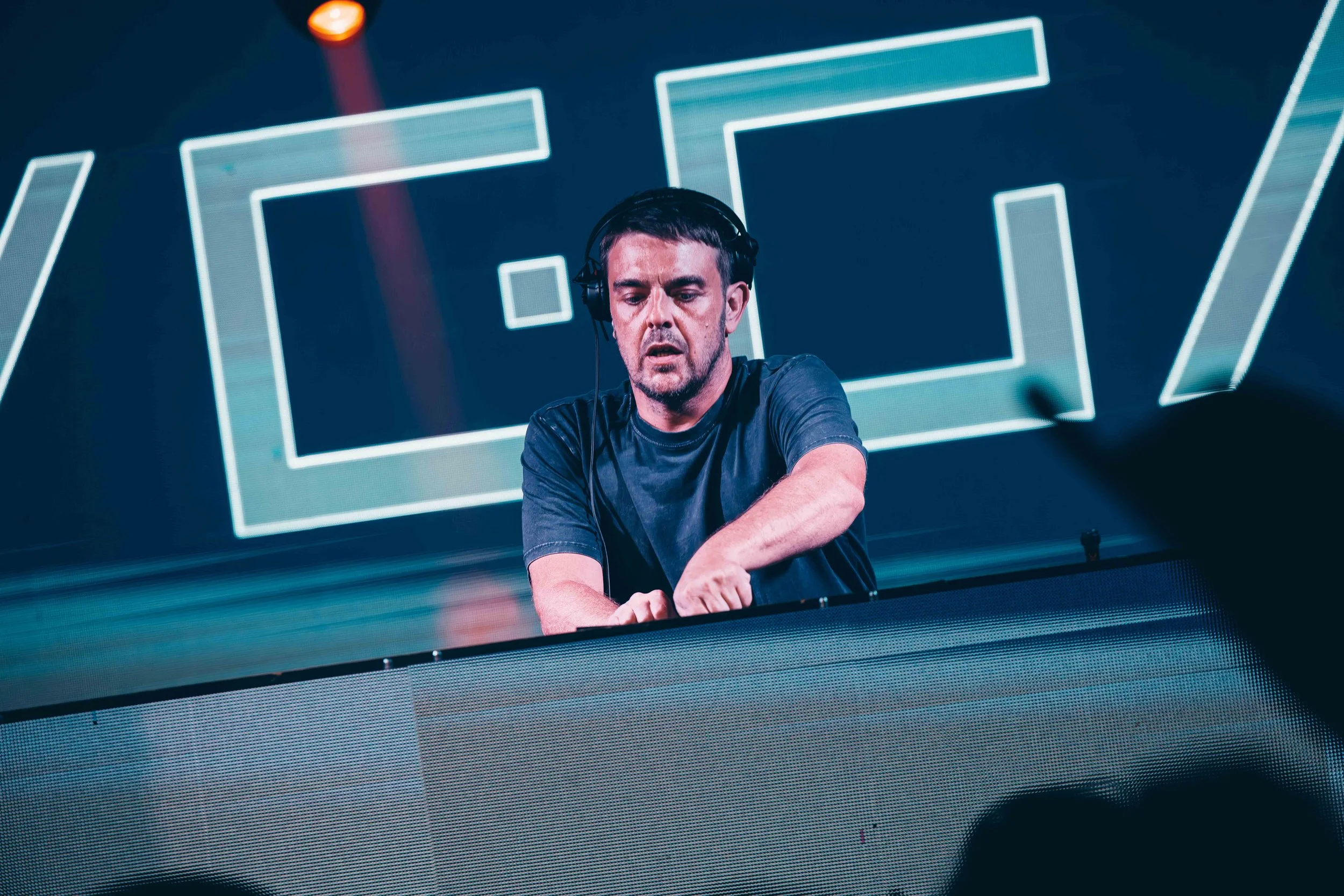 A male DJ with headphones playing music at a concert, with large LED screens in the background displaying geometric shapes.