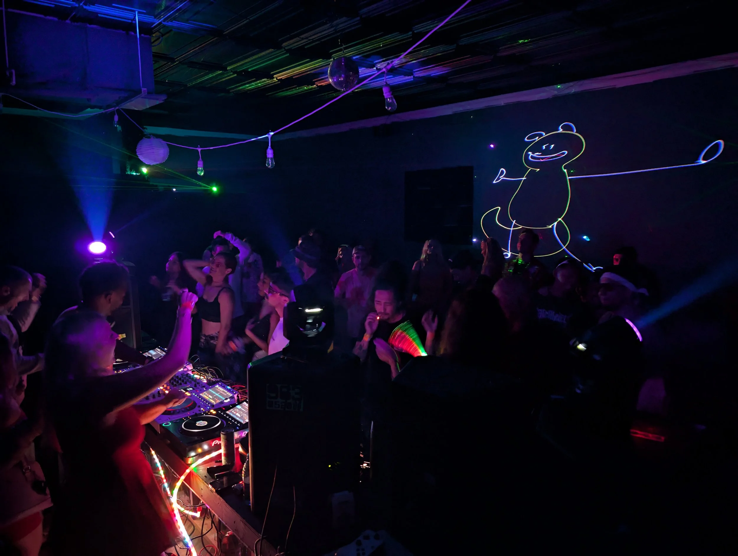 Dance floor with people dancing under colorful lights, DJ equipment, and neon decorations, including a cute animated character drawing on the wall.