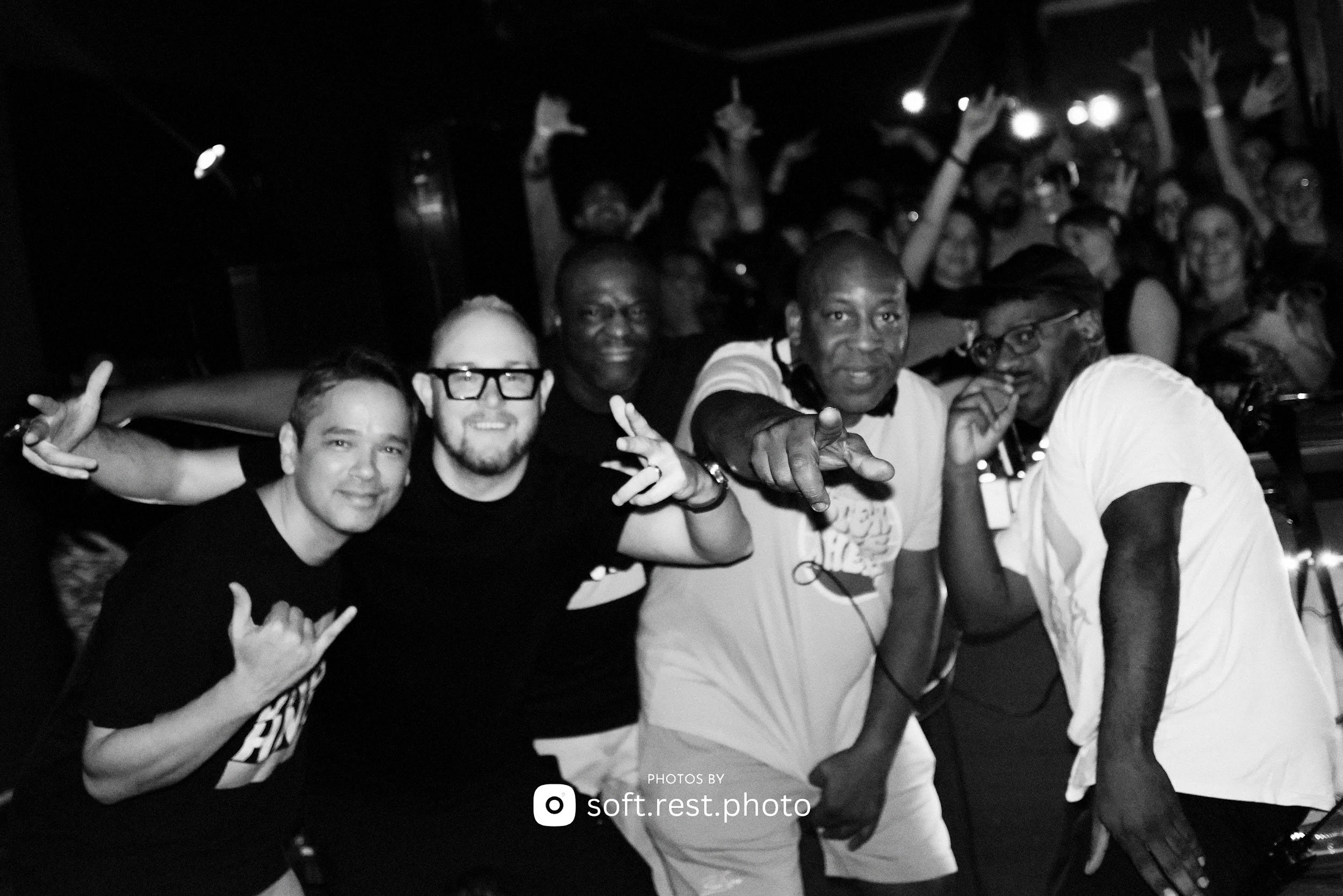 A group of five men in a club or concert setting, posing together and smiling at the camera, with a crowd of people dancing and enjoying music in the background.