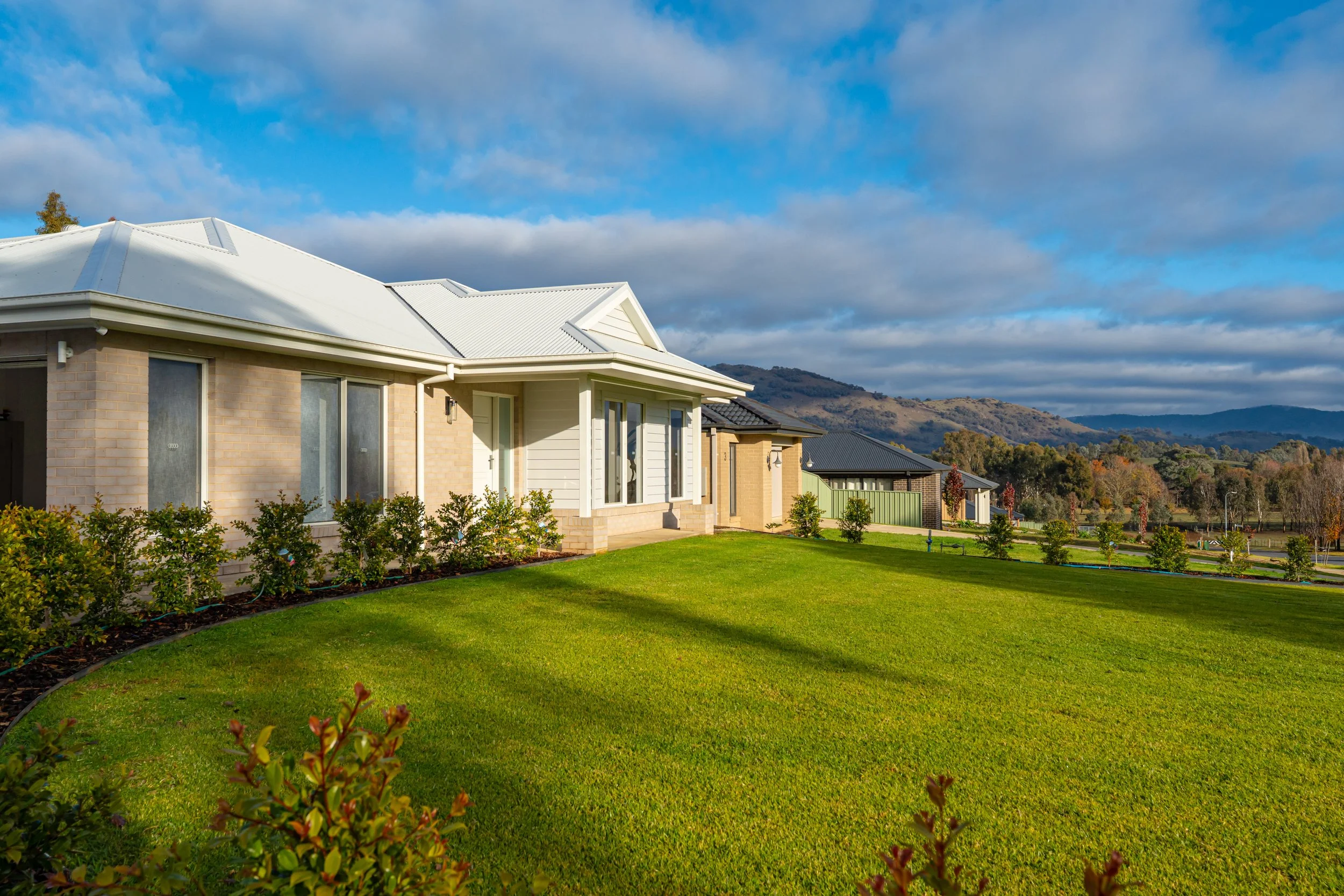 Living room with beige sofas, white chairs around a wooden dining table, mounted TV, ceiling fan, and decorative plants.