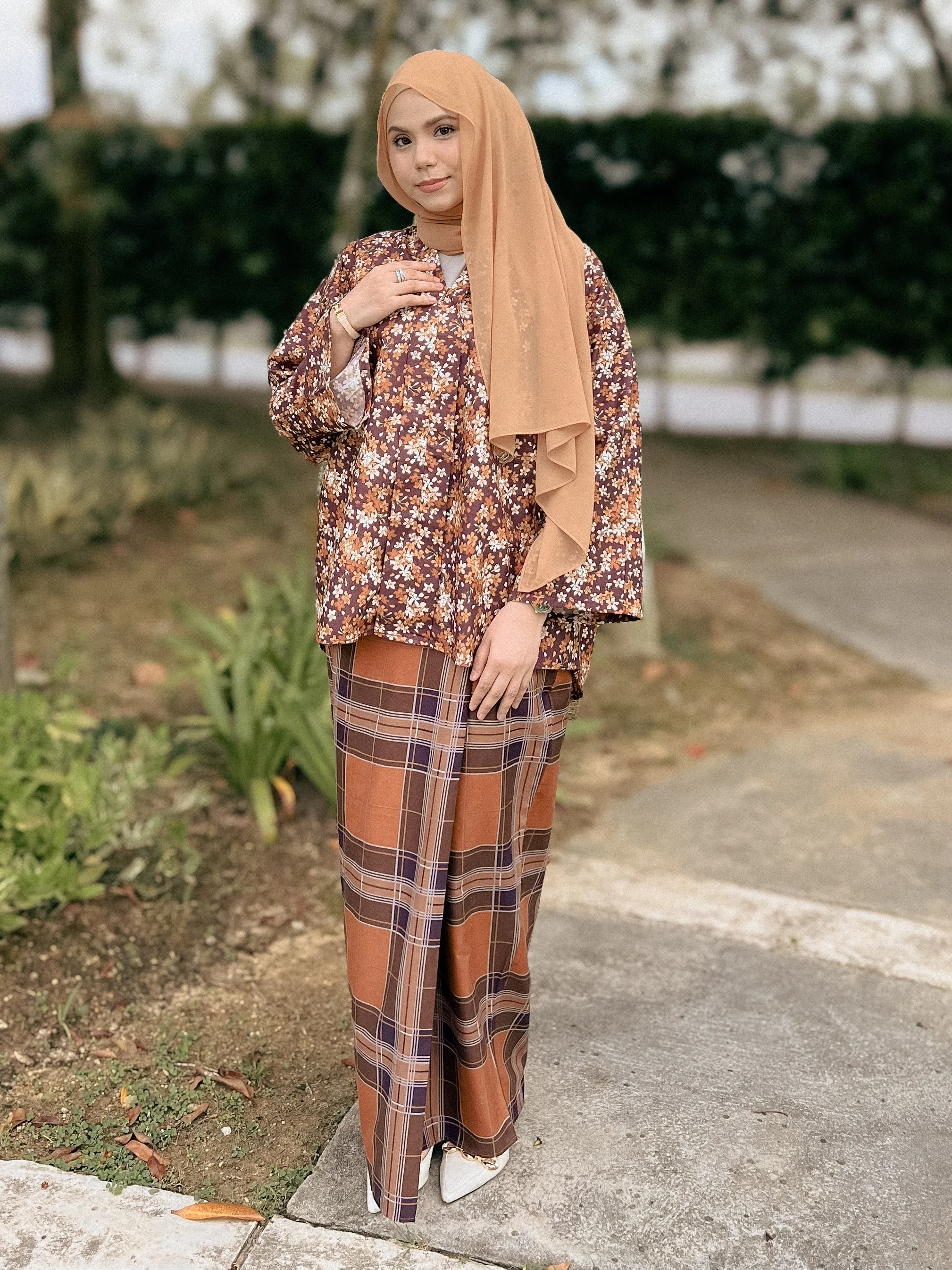 A woman wearing a tan hijab, a floral long-sleeve top, and plaid wide-leg pants standing on a sidewalk outdoors with greenery in the background.