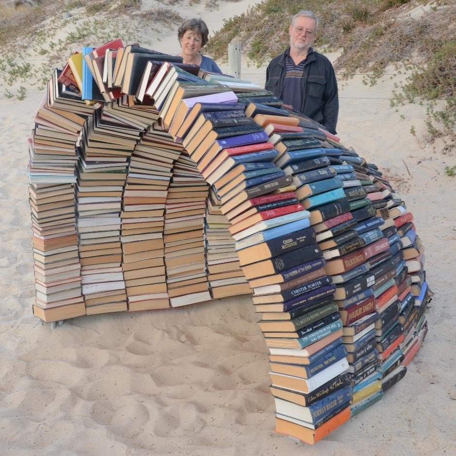 A few hundred books have been used to make a domed shelter on the beach. A man and woman, strangers, contemplate the beach sculpture.