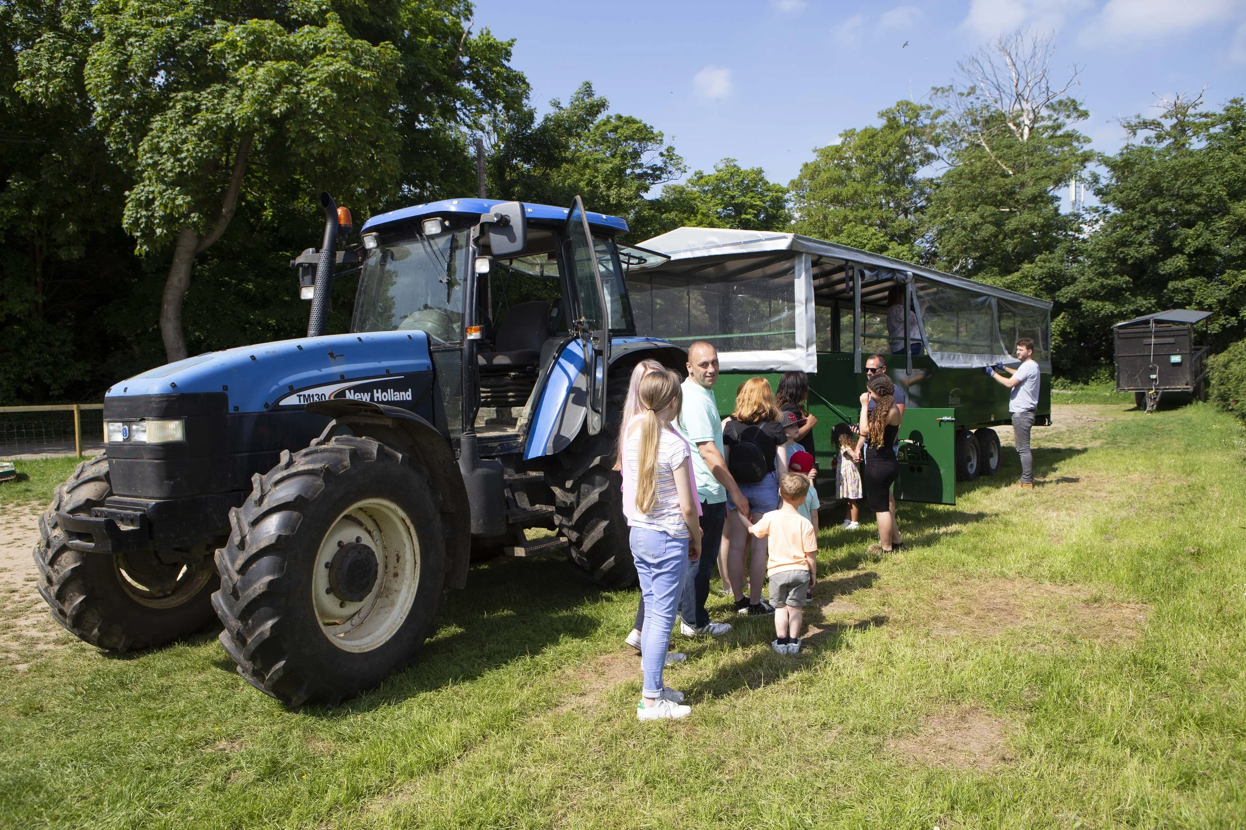People gather around a large blue tractor with an enclosed trailer attached, outdoors on a grassy area with trees in the background, on a sunny day.