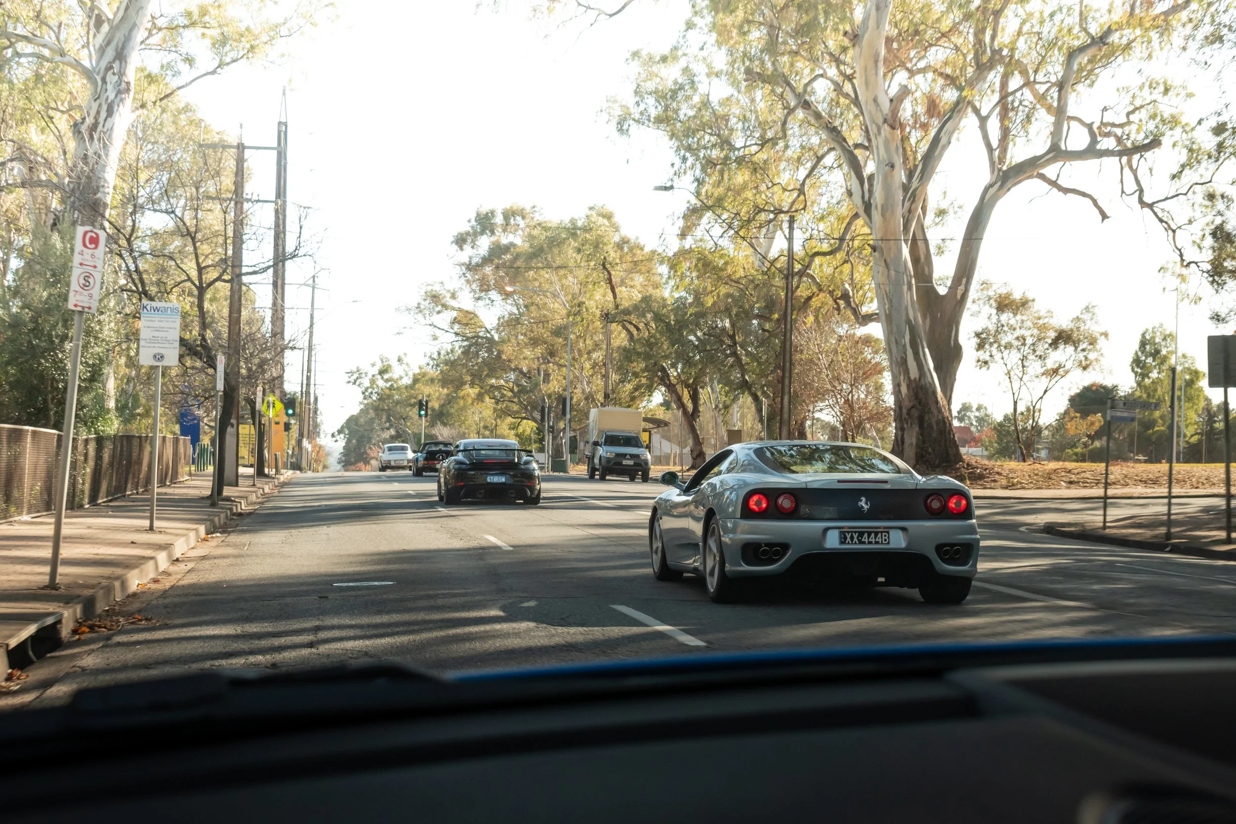 A street scene with a silver Ferrari car in the foreground and several other cars, including a black one and a white van, in the background. Large trees line the street, with some fall foliage, and streetlights and traffic signals are visible.