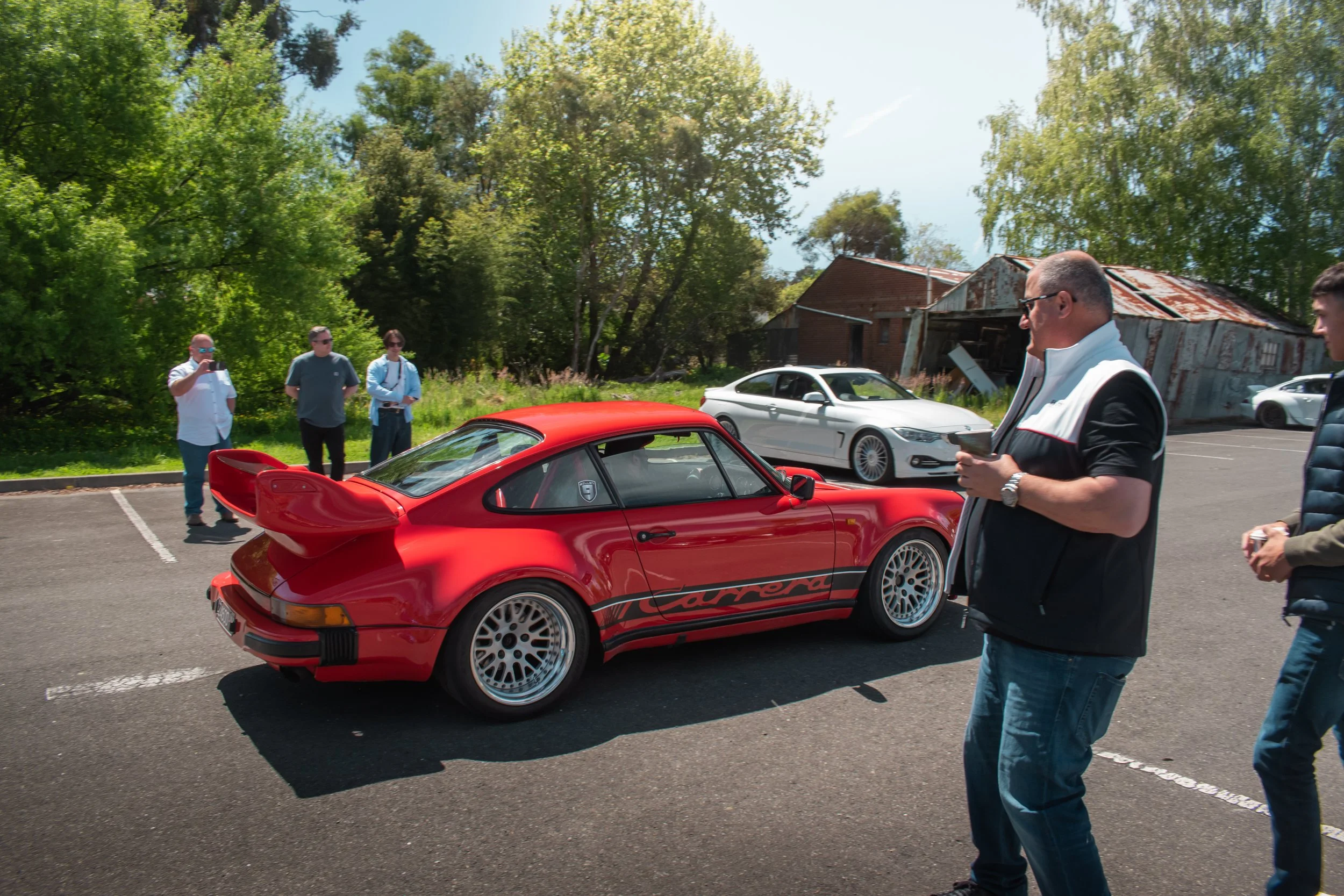 Red classic Porsche Carrera sports car parked in an outdoor lot with five people around, and trees and dilapidated buildings in the background.