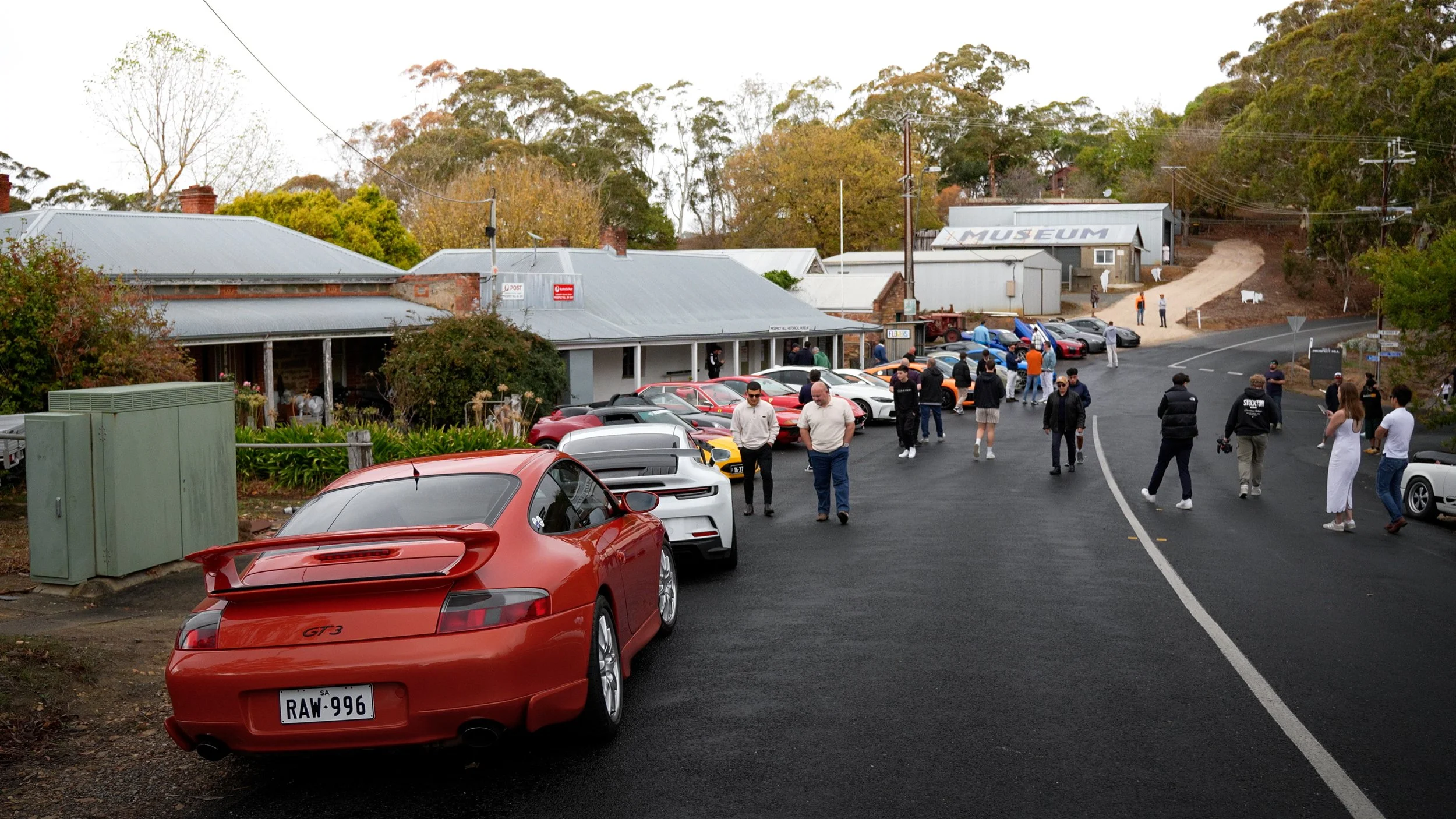 A group of people gathered outside a museum with classic and sports cars parked along the side of the road, including a red Porsche 911 GT3, with trees and a dirt road leading uphill in the background.