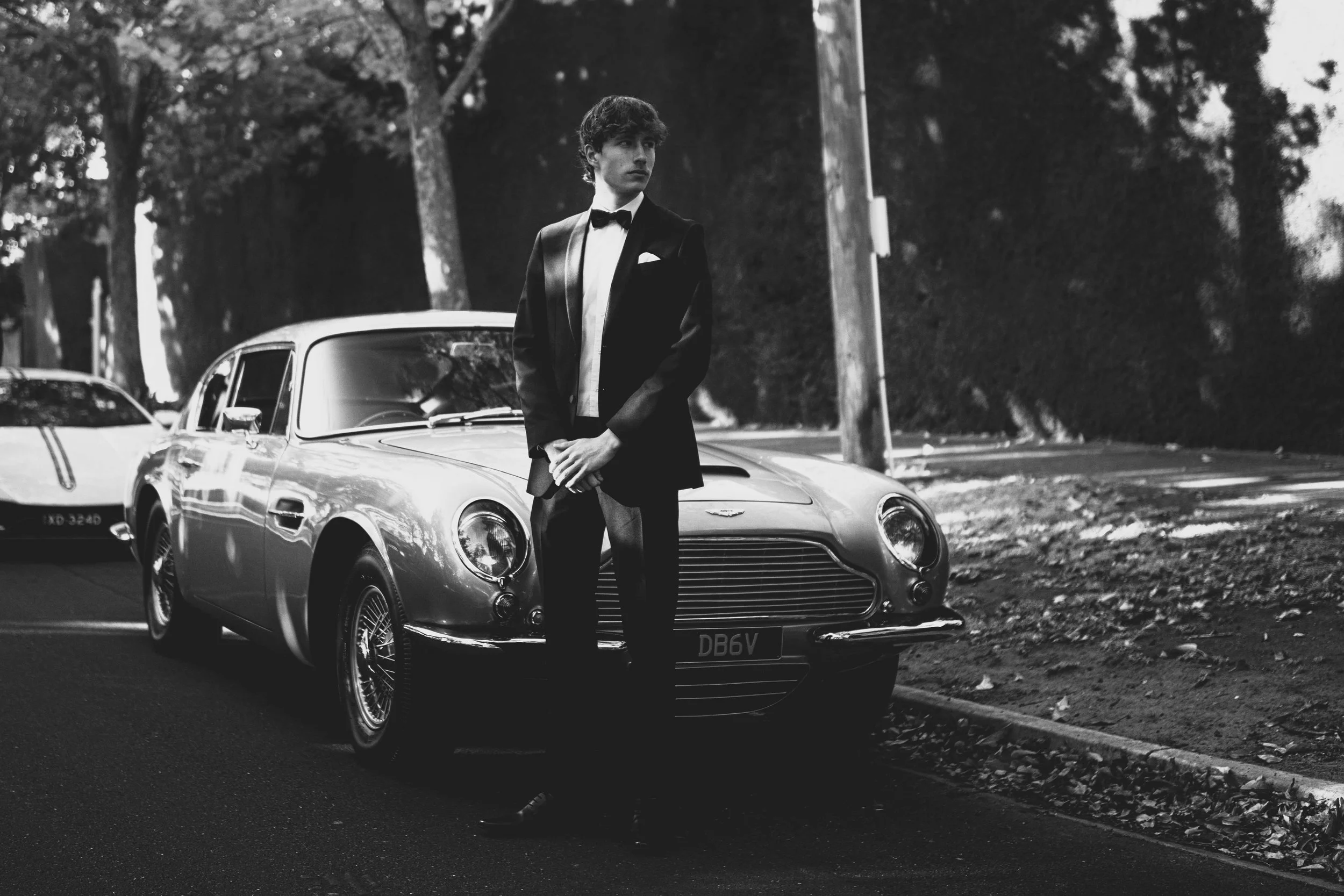 A young man in a tuxedo standing in front of a vintage sports car on a street, black and white photo