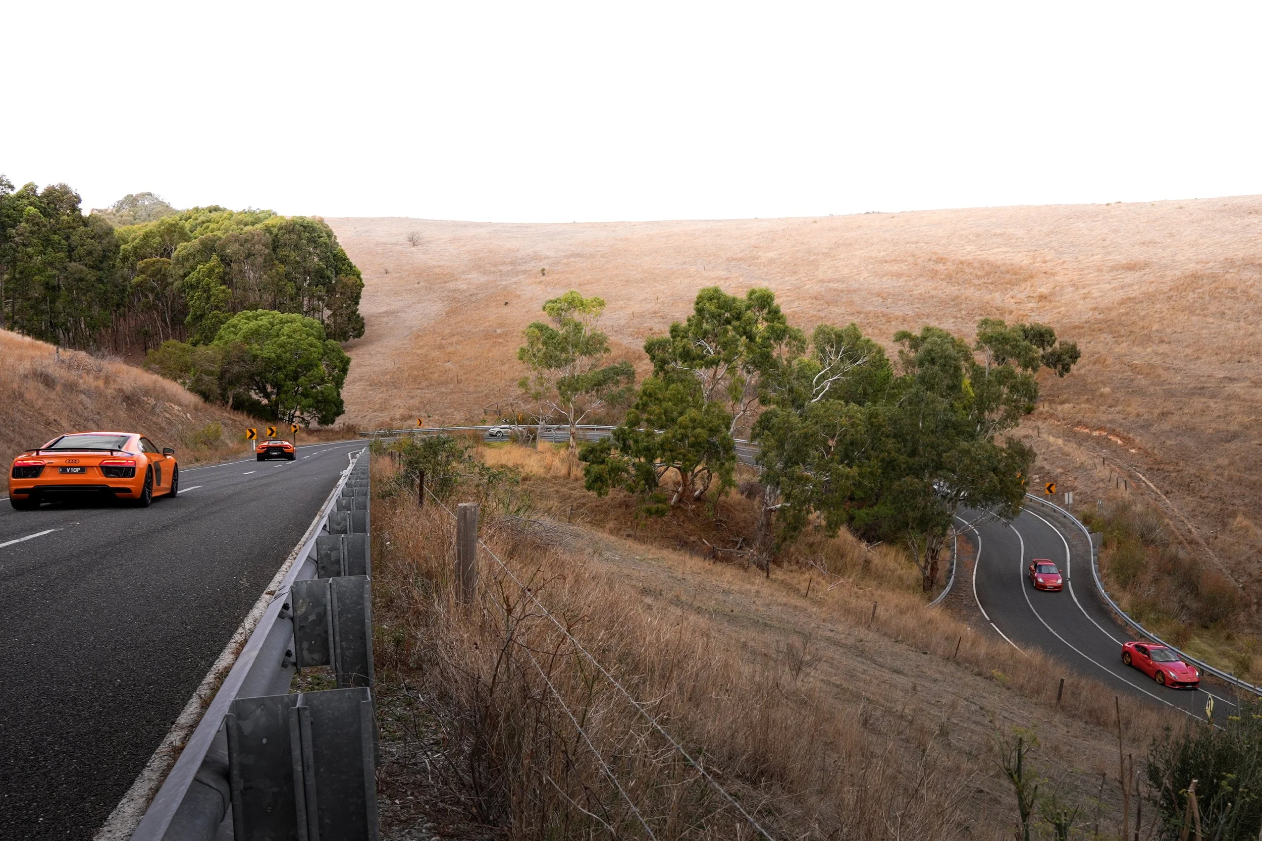 Two orange sports cars on a winding mountain road, with a red police car further ahead. The road is bordered by dry grassy hills and green trees.