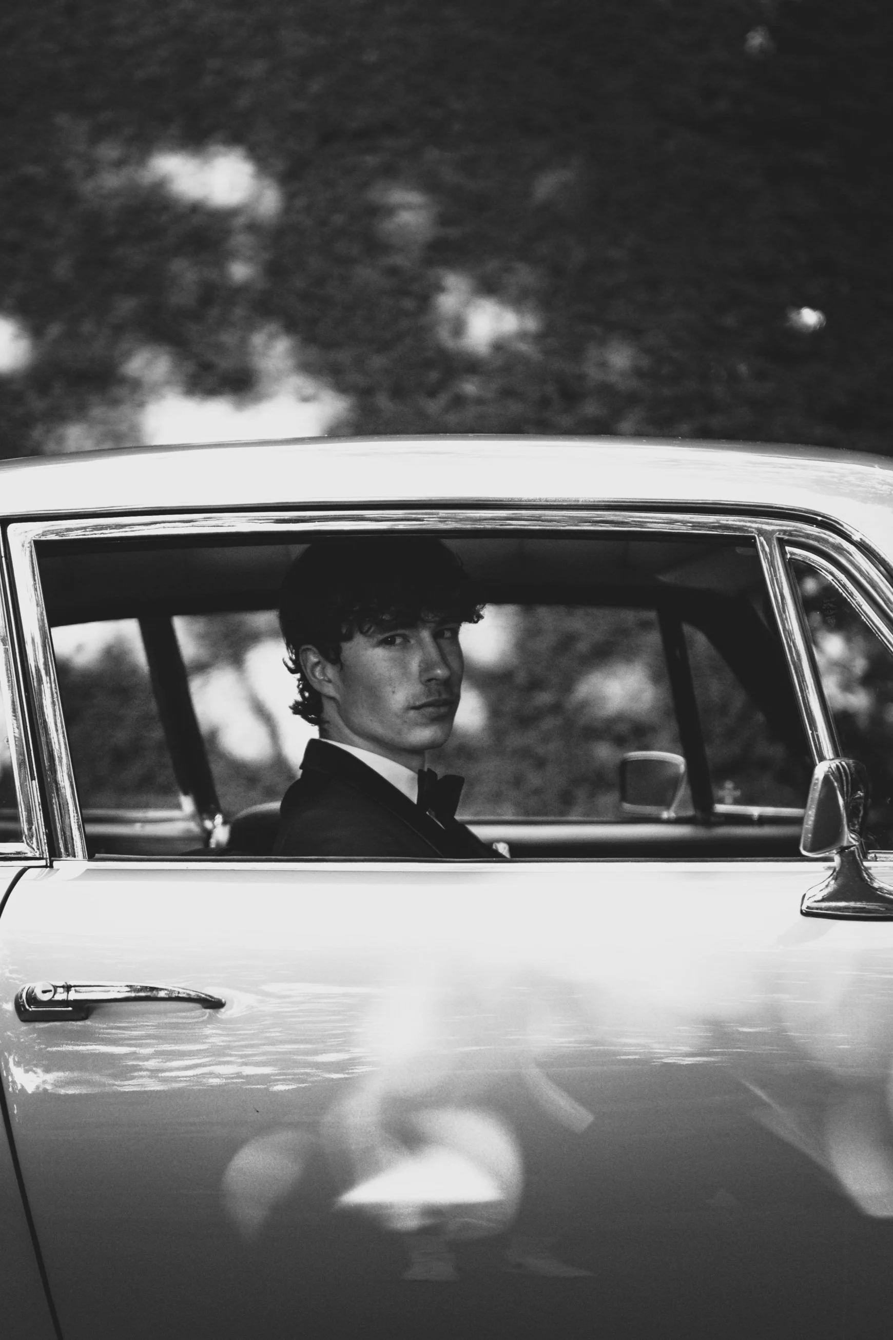 Black and white photo of a young man in a tuxedo sitting in the back seat of a vintage car, looking towards the camera with a serious expression, with trees and clouds in the background.