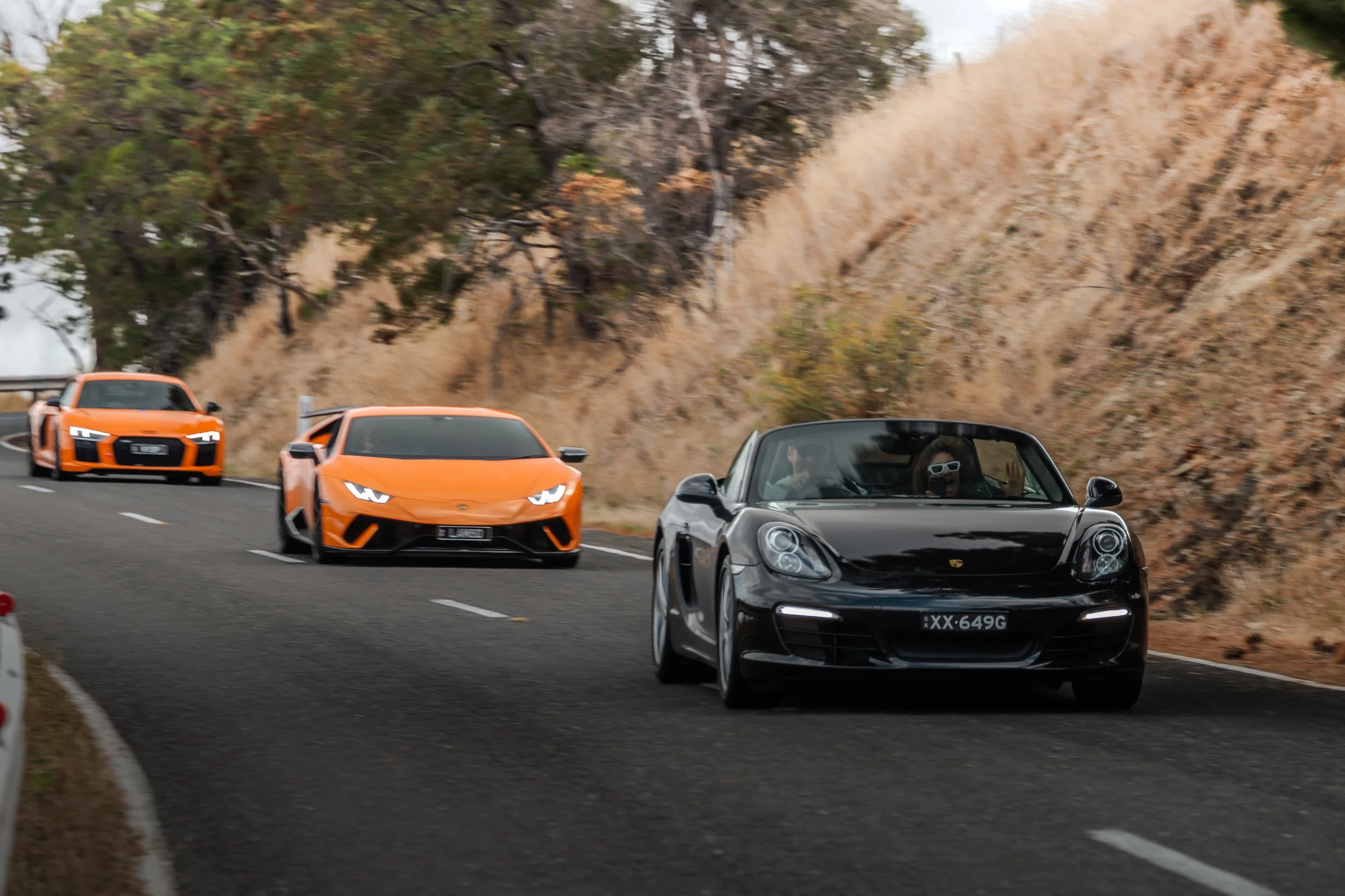 A black Porsche convertible leading a convoy of three orange Lamborghini sports cars on a winding mountain road with dry, rocky terrain and trees in the background.
