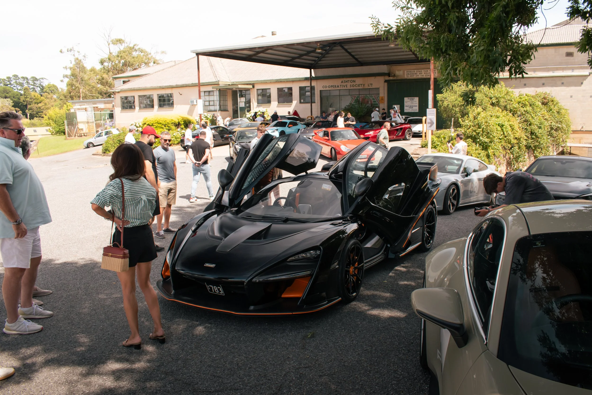 A black supercar with scissor doors open at a car show, surrounded by people and other exotic cars, in front of a building with a sign that reads 'Ashton Cooperative Society'.