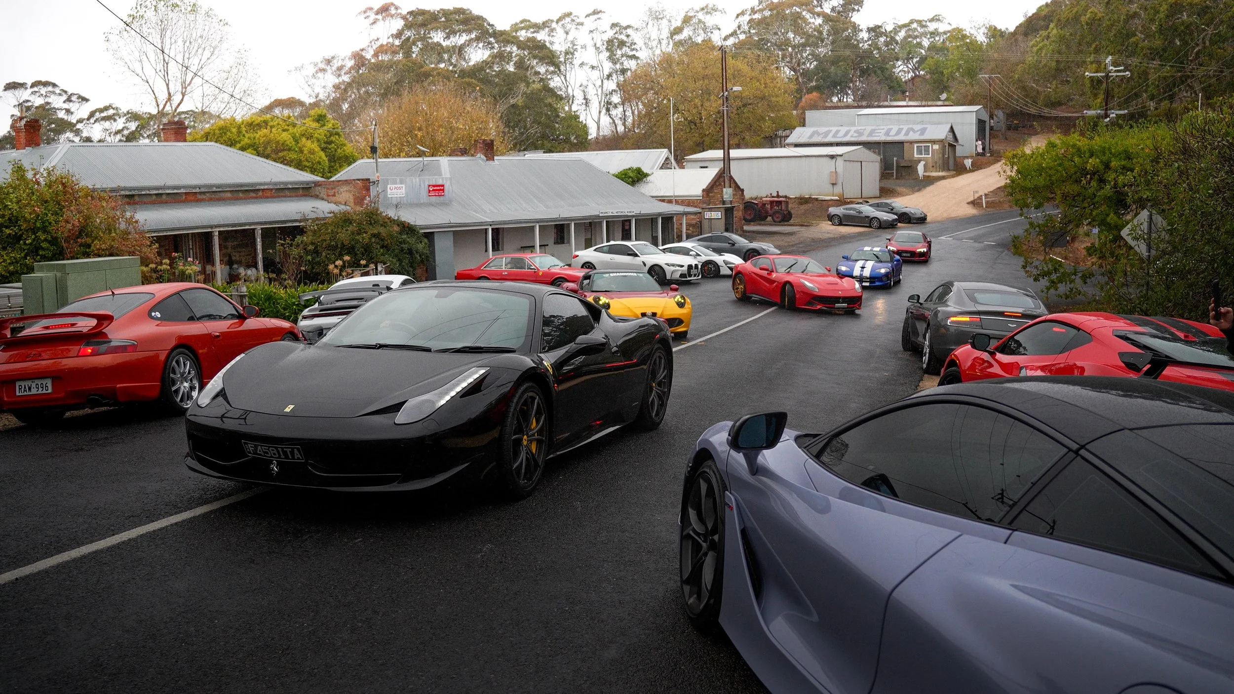 A collection of luxury sports cars, including Ferraris and other high-end models, parked outside a building with a museum sign on the hill in the background.
