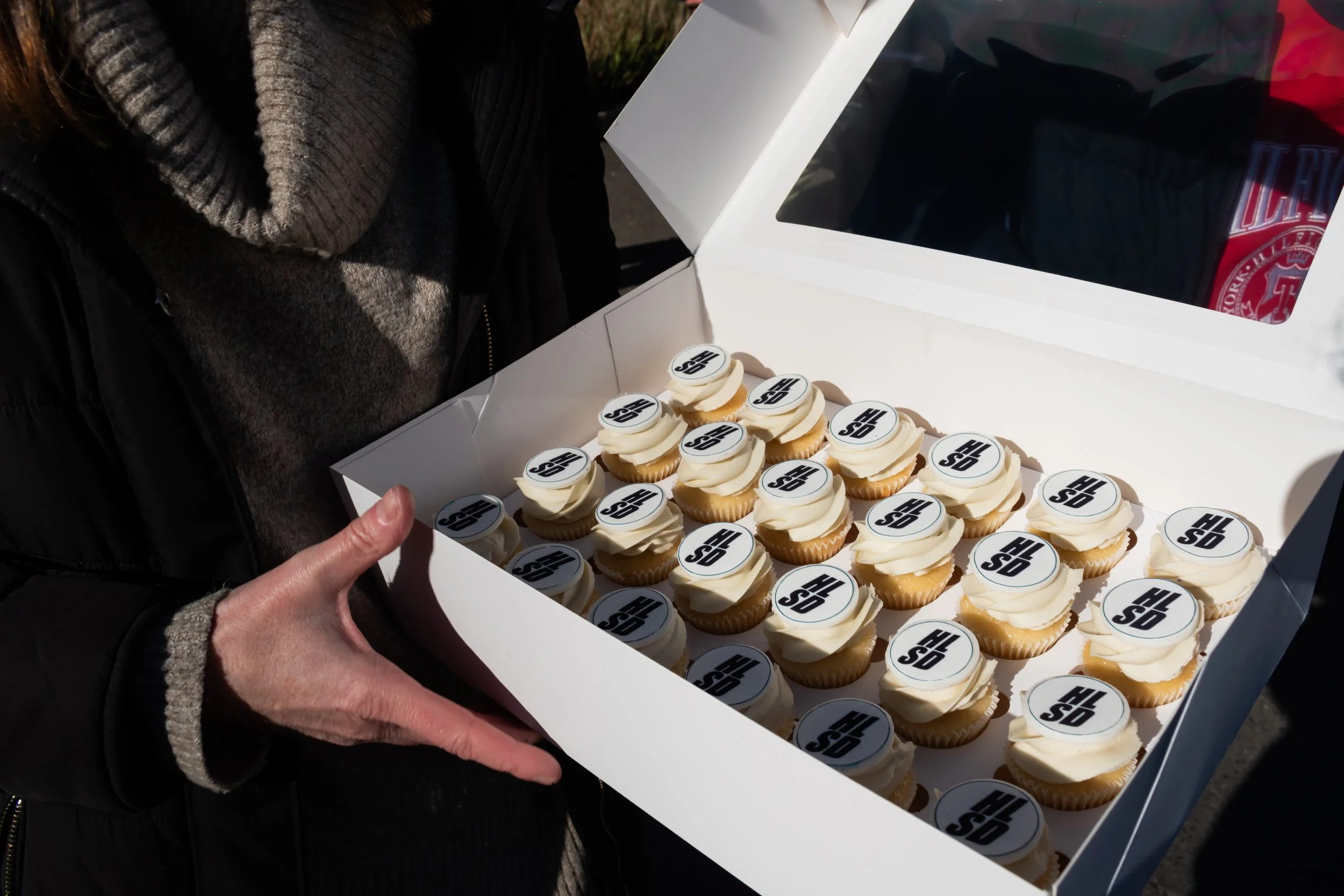 Person holding a box of cupcakes decorated with logos on top.