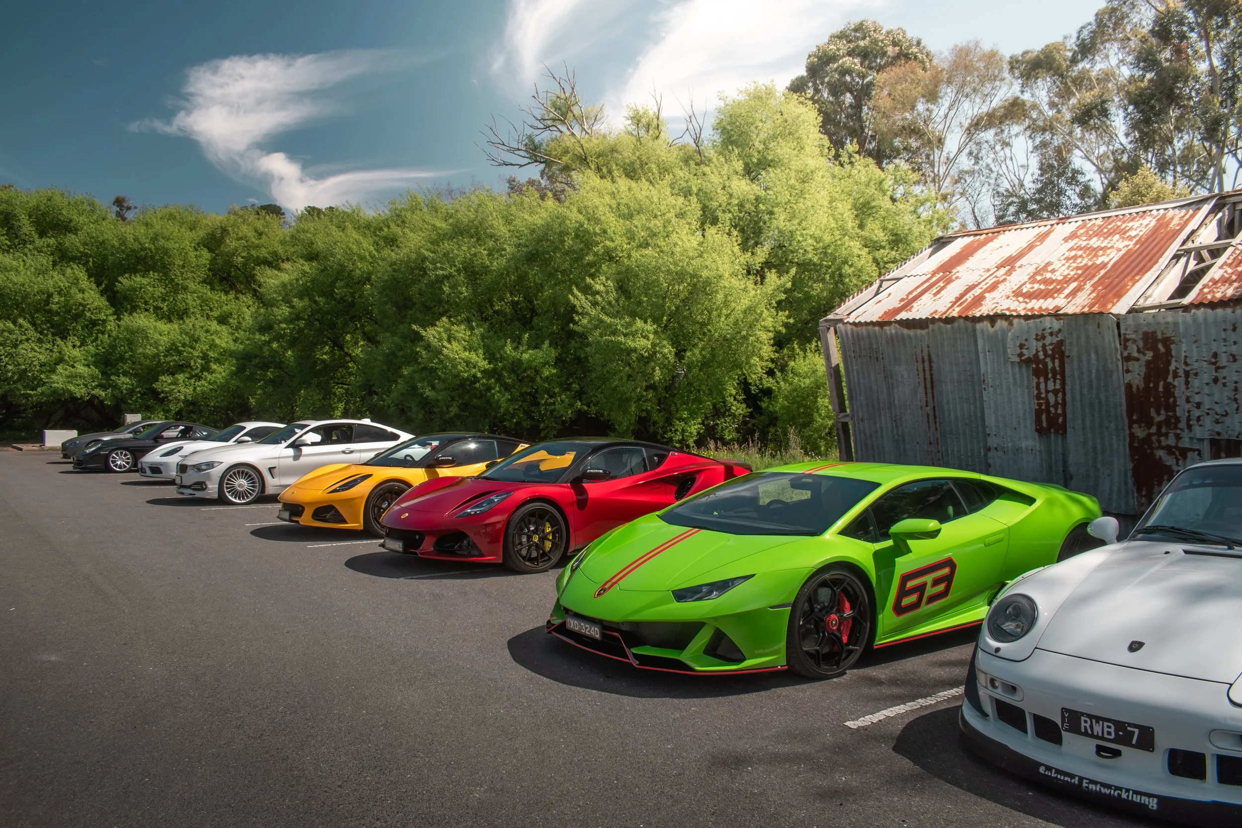 A lineup of luxury sports cars parked on an asphalt surface with green trees and a weathered metal shed in the background.