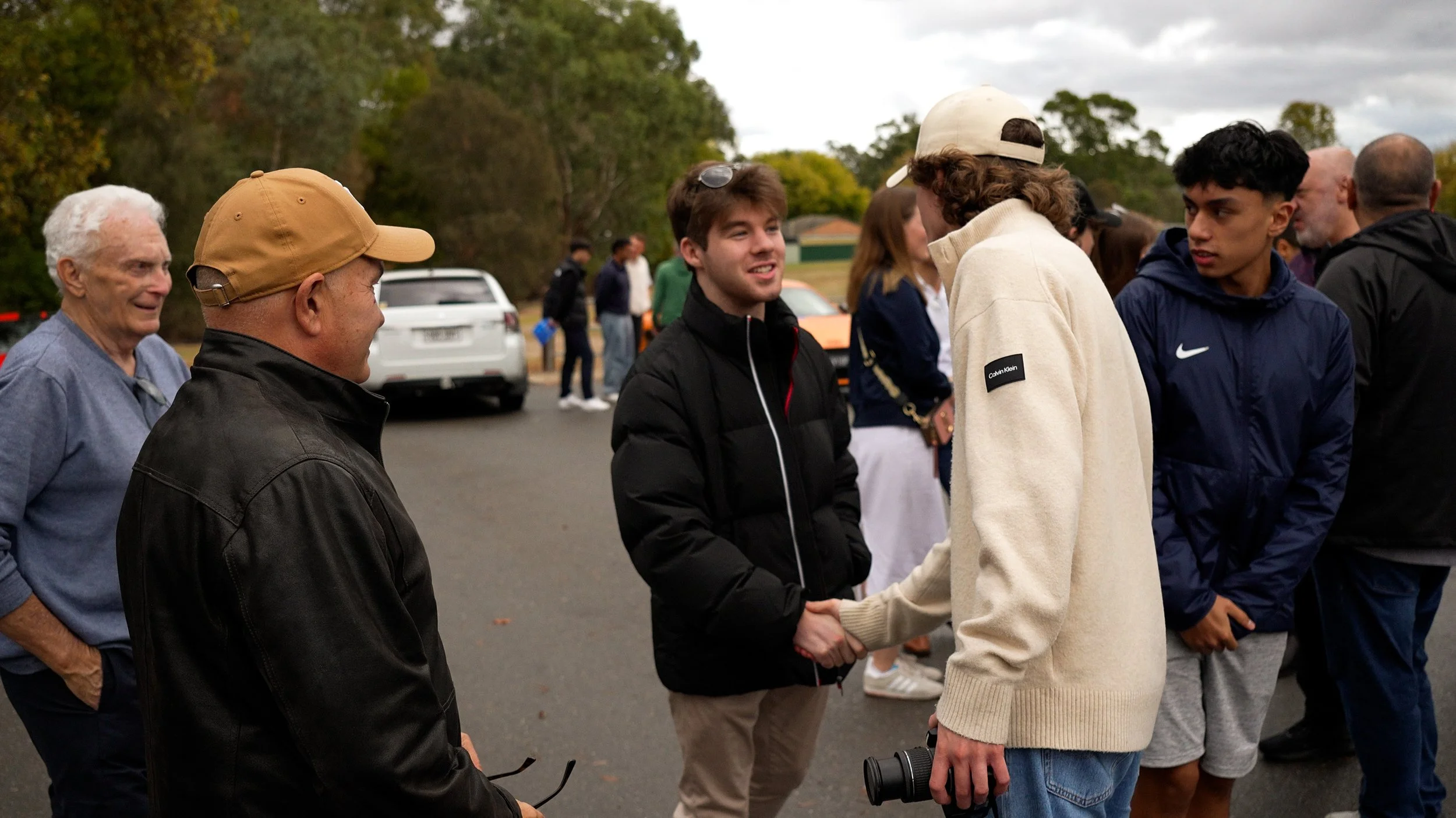 Young man shaking hands with older man outdoors, other people in line, cars in background, trees, cloudy sky.