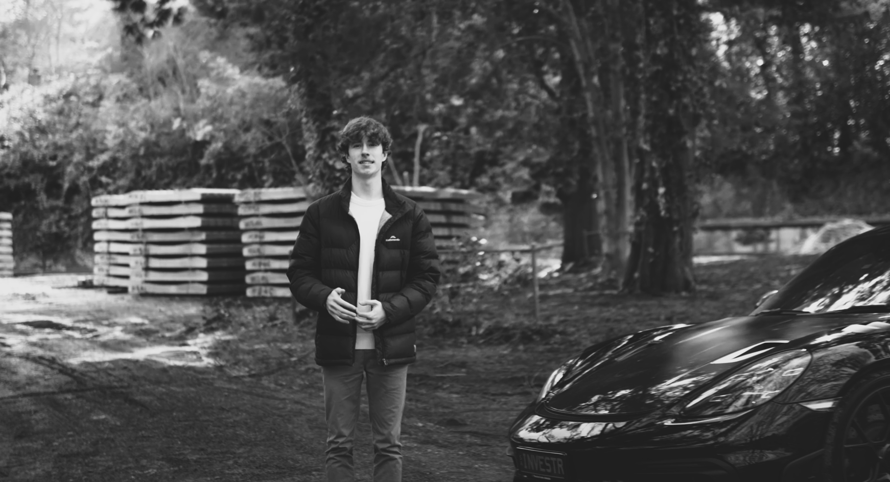 A young man with curly hair standing outdoors next to a sleek black sports car, holding a coffee cup, with trees and stacked wood in the background.