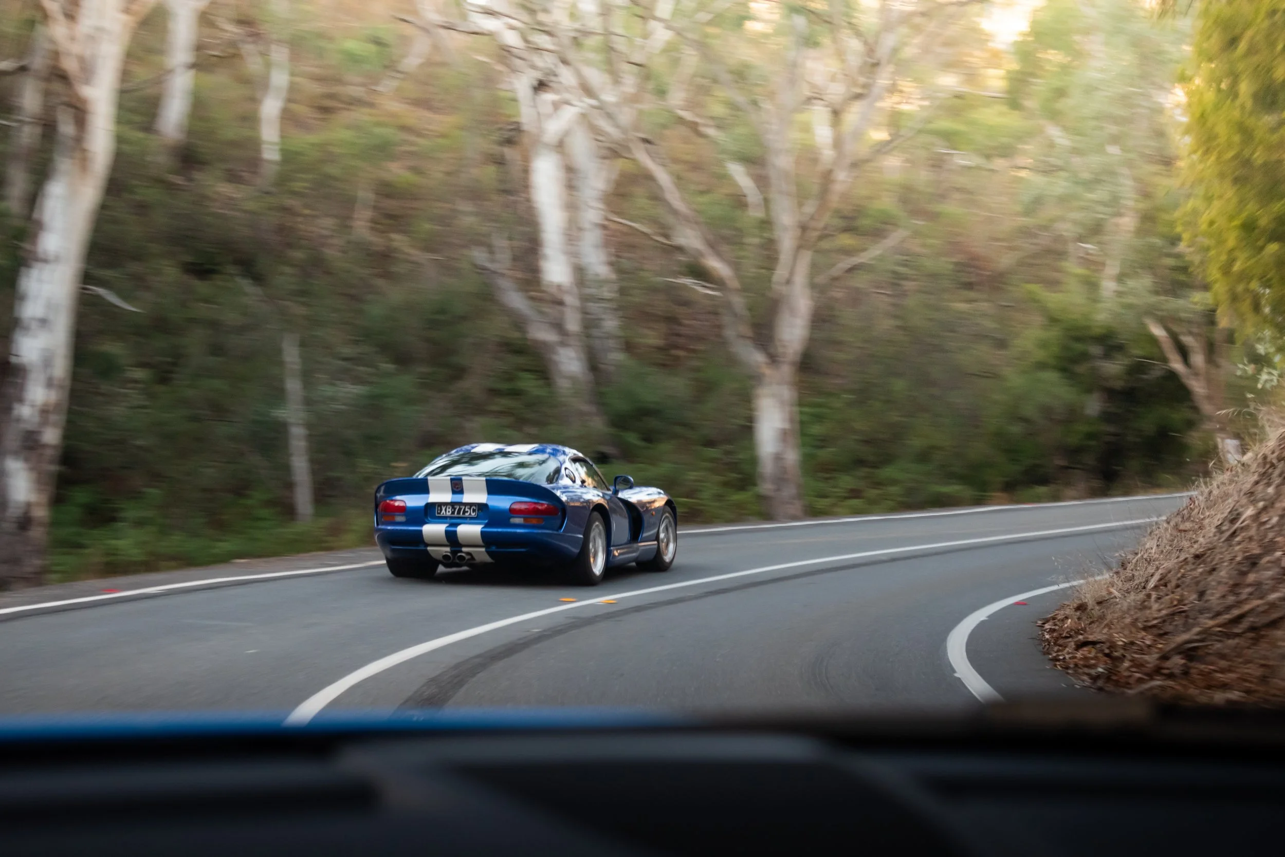 A blue sports car with white racing stripes driving around a sharp turn on a winding mountain road lined with trees.