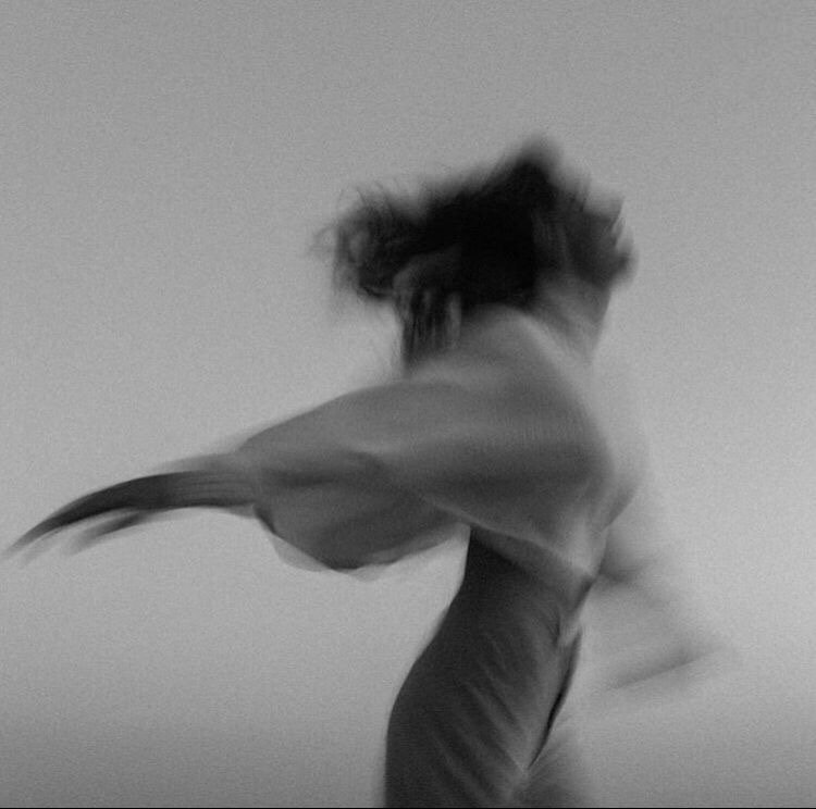 A person with tousled hair spinning or dancing, captured mid-movement against a plain wall, in black and white.