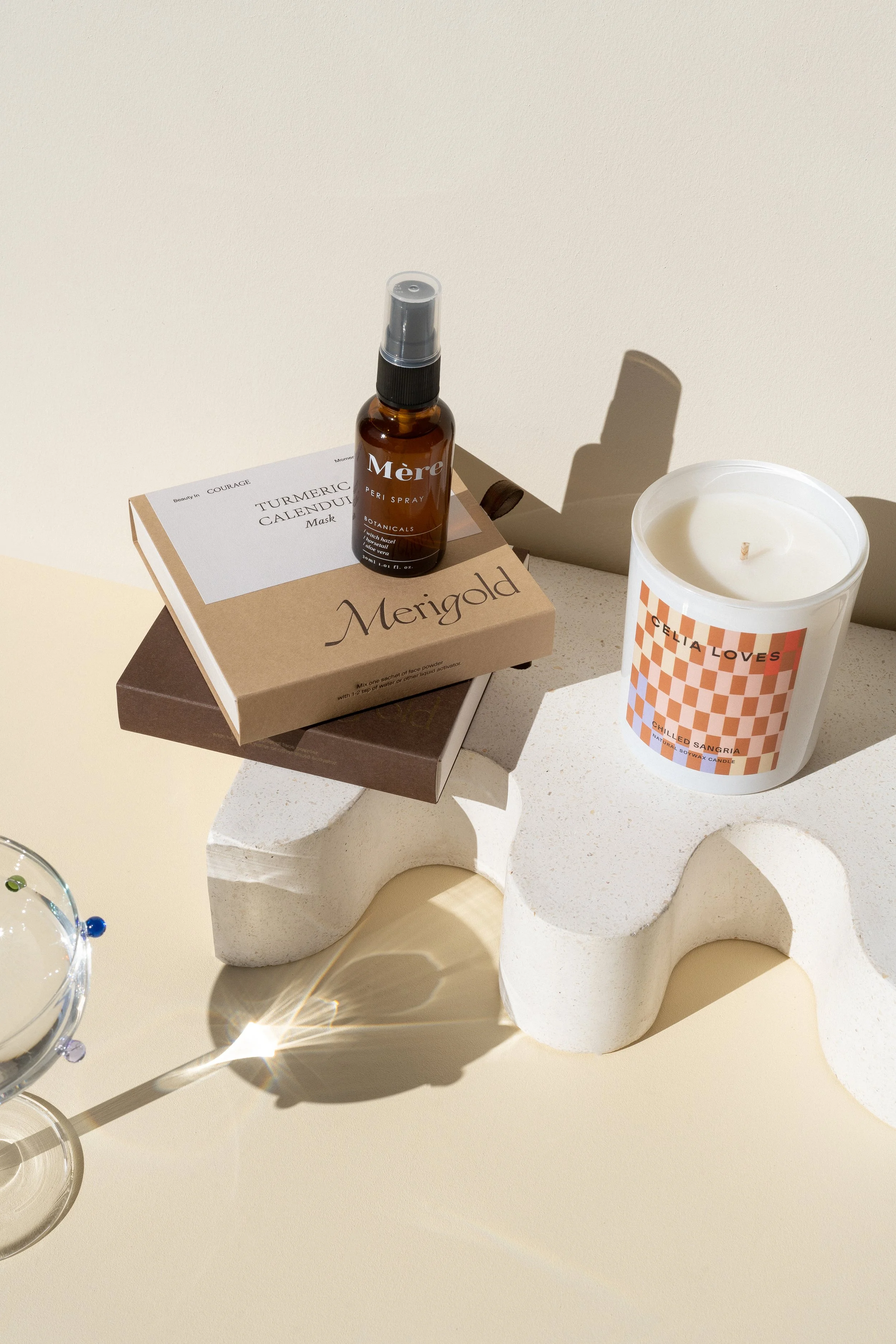 A white candle labeled 'Celia Loves Chilled Sangria' on a textured white abstract sculpture, with brown and beige boxes and a small spray bottle of 'Mère' botanical peri spray on top.